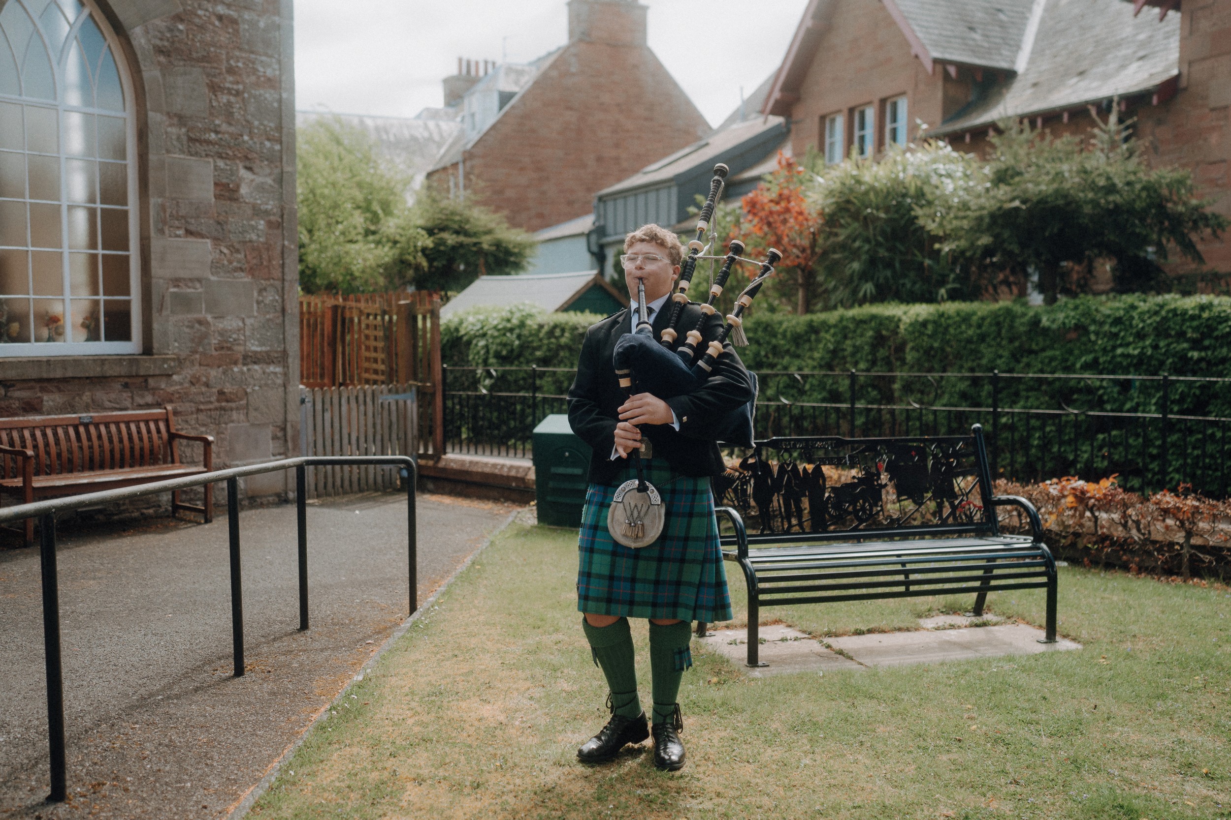 A person in traditional Scottish attire stands on grass playing bagpipes in front of a building with stone walls and surrounding greenery.