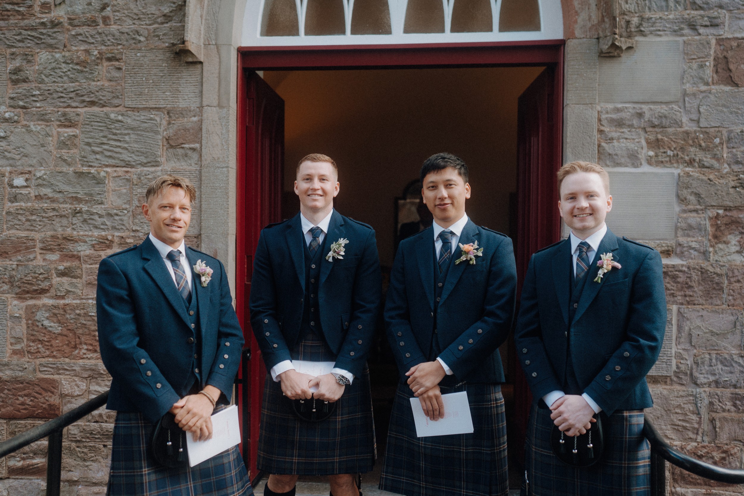Four men dressed in matching kilts and jackets stand in front of a stone building entrance, smiling and holding papers.