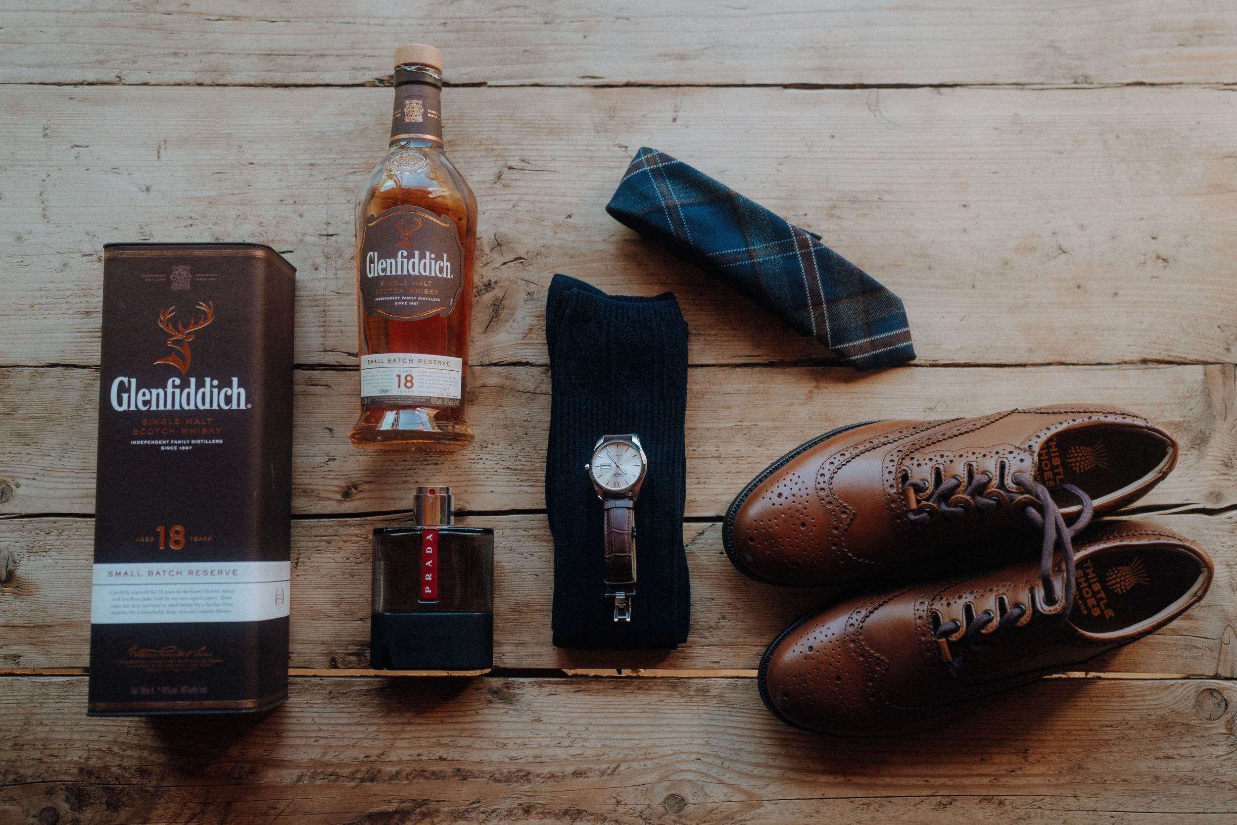 A bottle of Glenfiddich 18, its box, cologne, a wristwatch, blue socks, a plaid tie, and brown dress shoes arranged on a wooden floor.