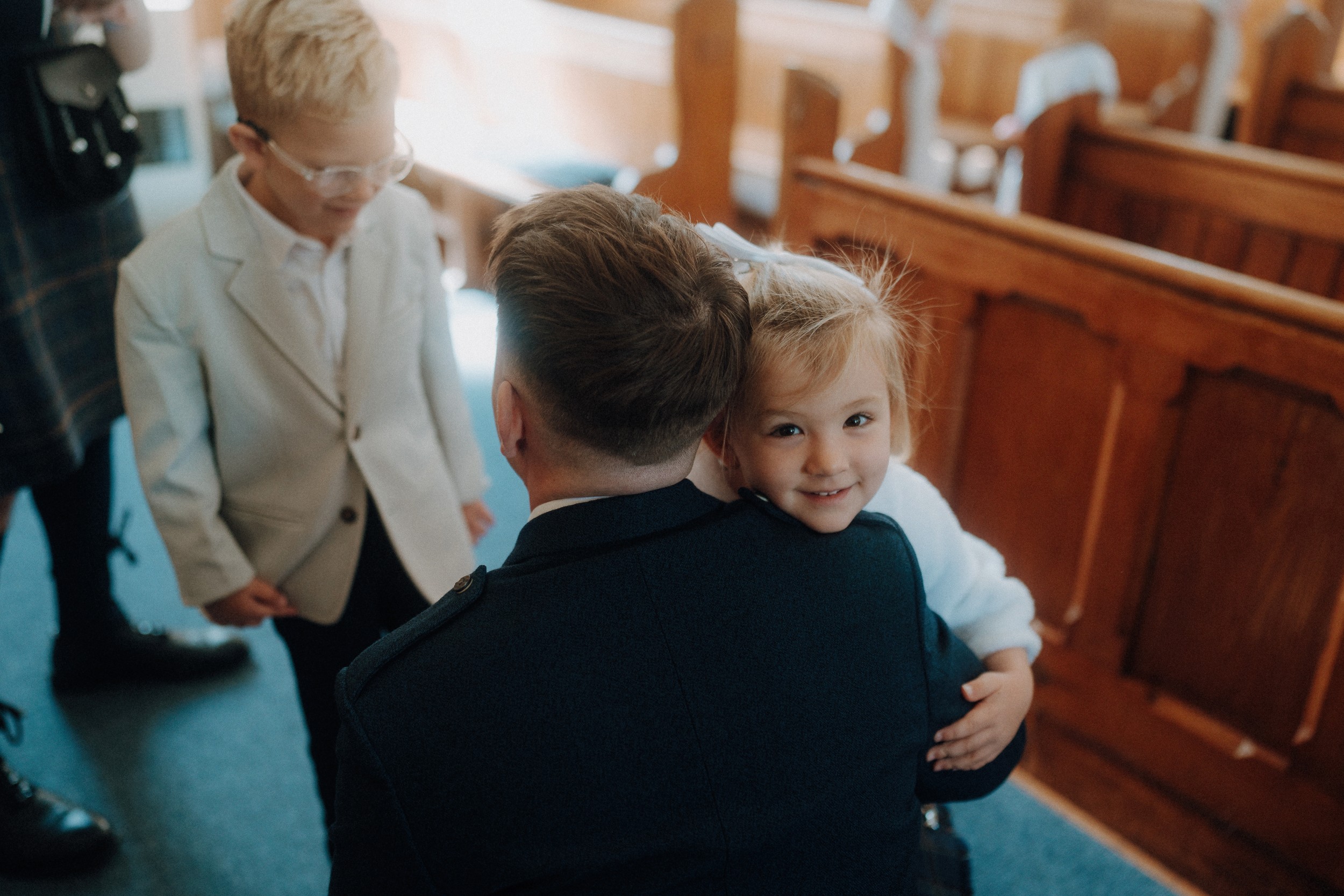 A young girl hugs an adult dressed formally inside a wooden pew-filled room, while a boy in glasses and a light suit stands nearby.