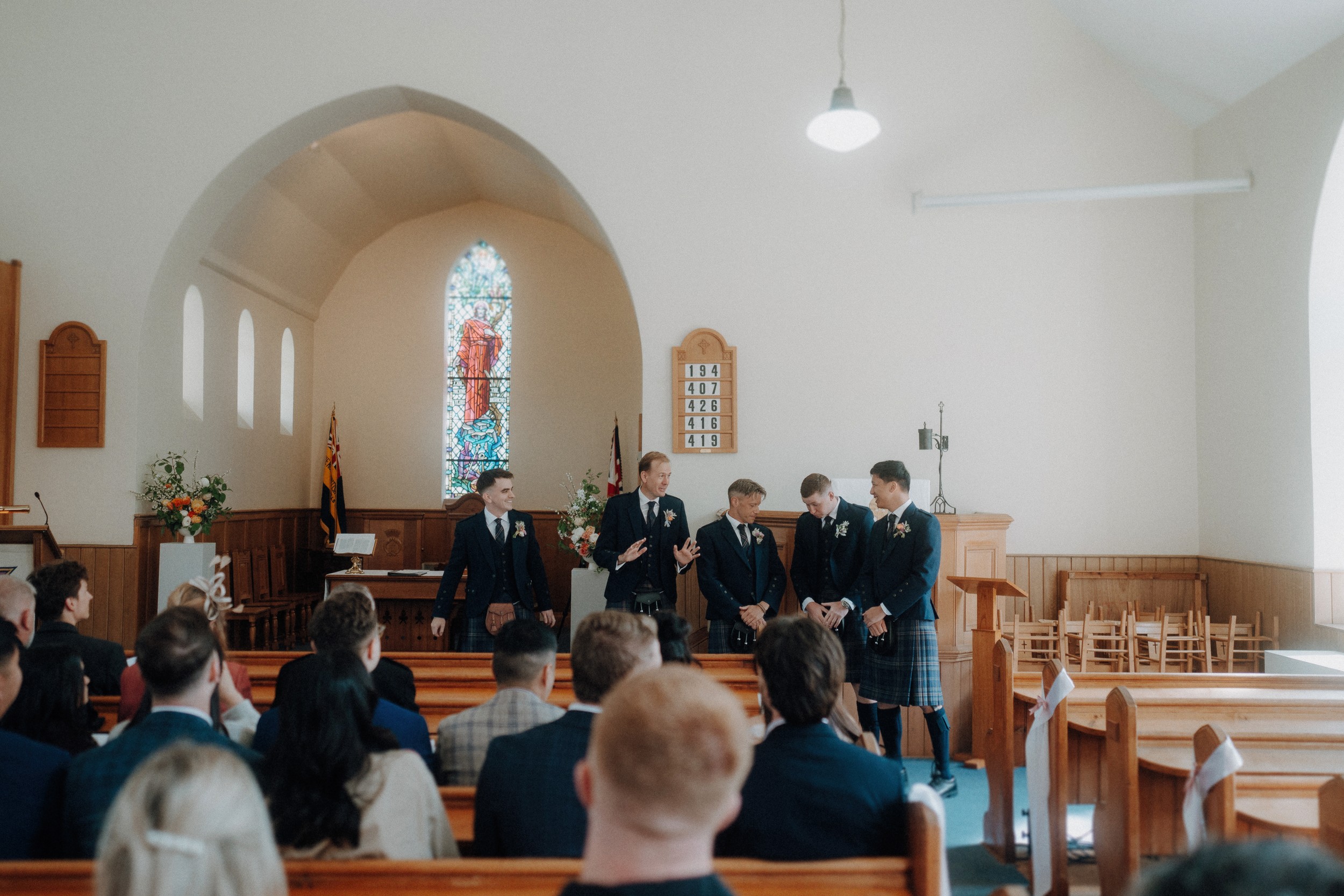Five men in suits stand at the front of a church with wooden pews, while guests seated in the foreground watch. A stained glass window is visible behind them.