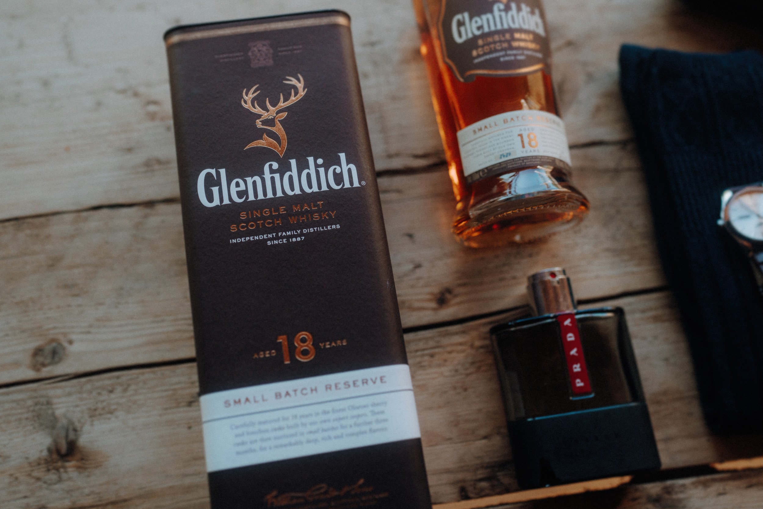 A Glenfiddich 18 Year Old Scotch whisky box and bottle on a wooden surface next to a Prada cologne bottle and a partial view of a watch and dark fabric.