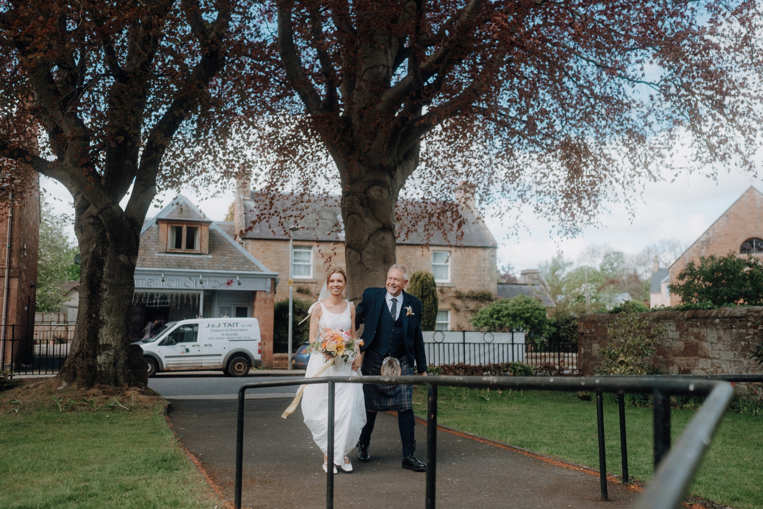 A bride in a white dress walks outside on a path beside a man in a suit and kilt, both smiling, with trees and houses in the background.