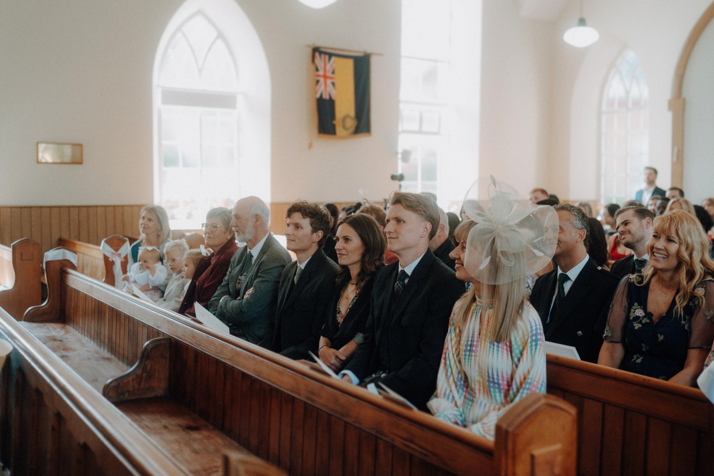 A group of people dressed formally sit in wooden pews inside a church during a daytime ceremony, with natural light coming through arched windows.