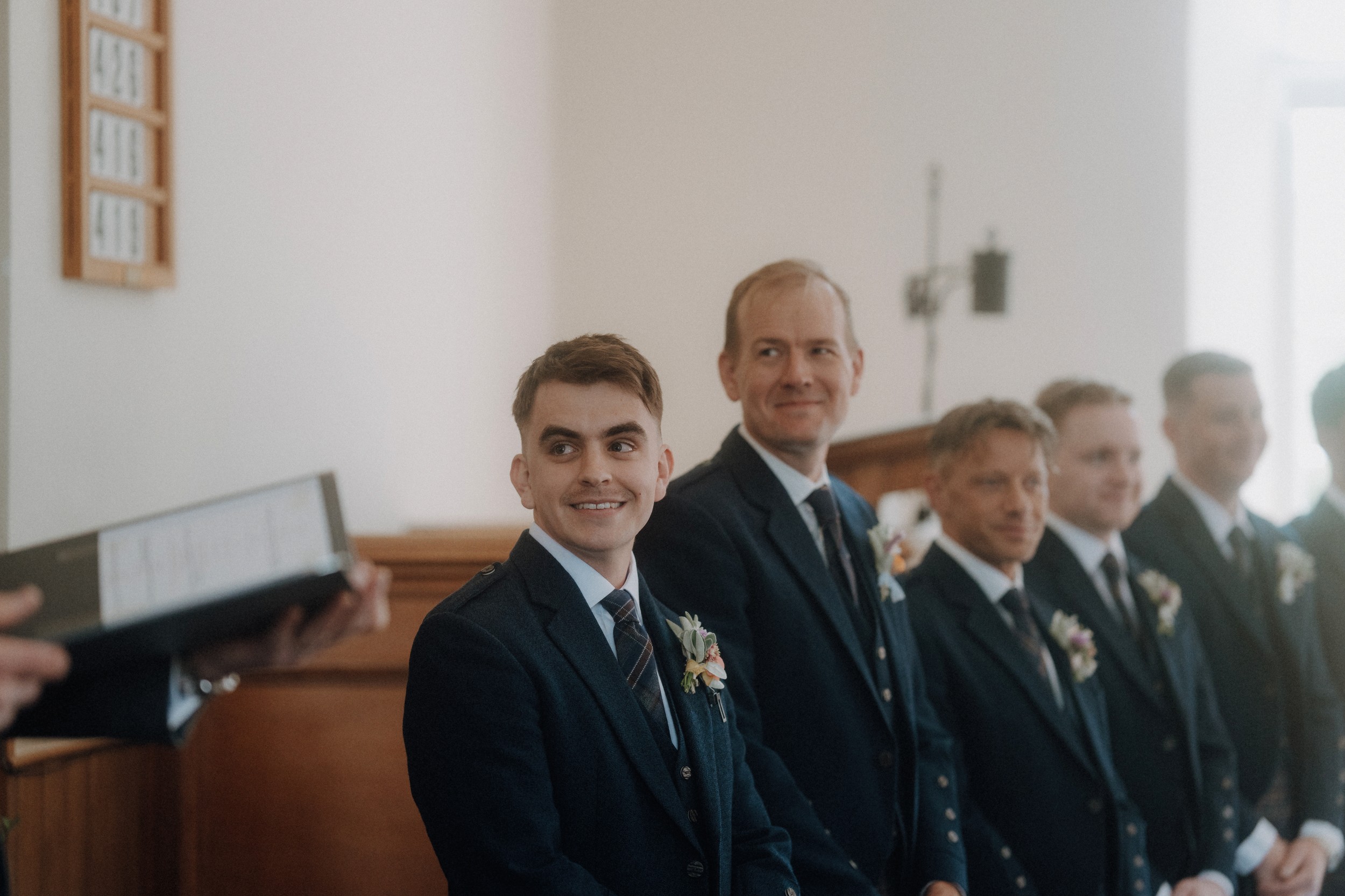 A group of men in suits with boutonnieres stand in a line at the front of a room, smiling and facing a person holding a binder.