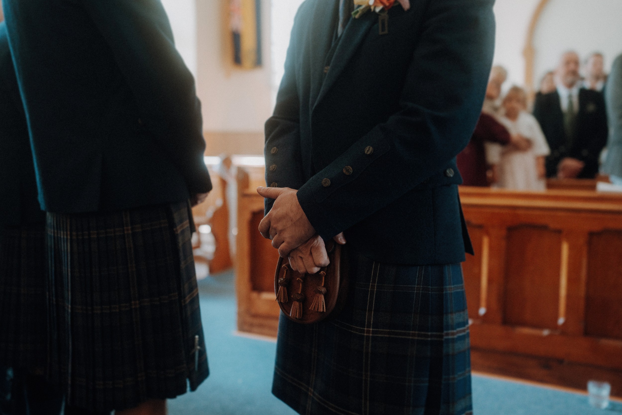 Two people wearing traditional Scottish kilts stand inside a church, with hands clasped and pews visible in the background.
