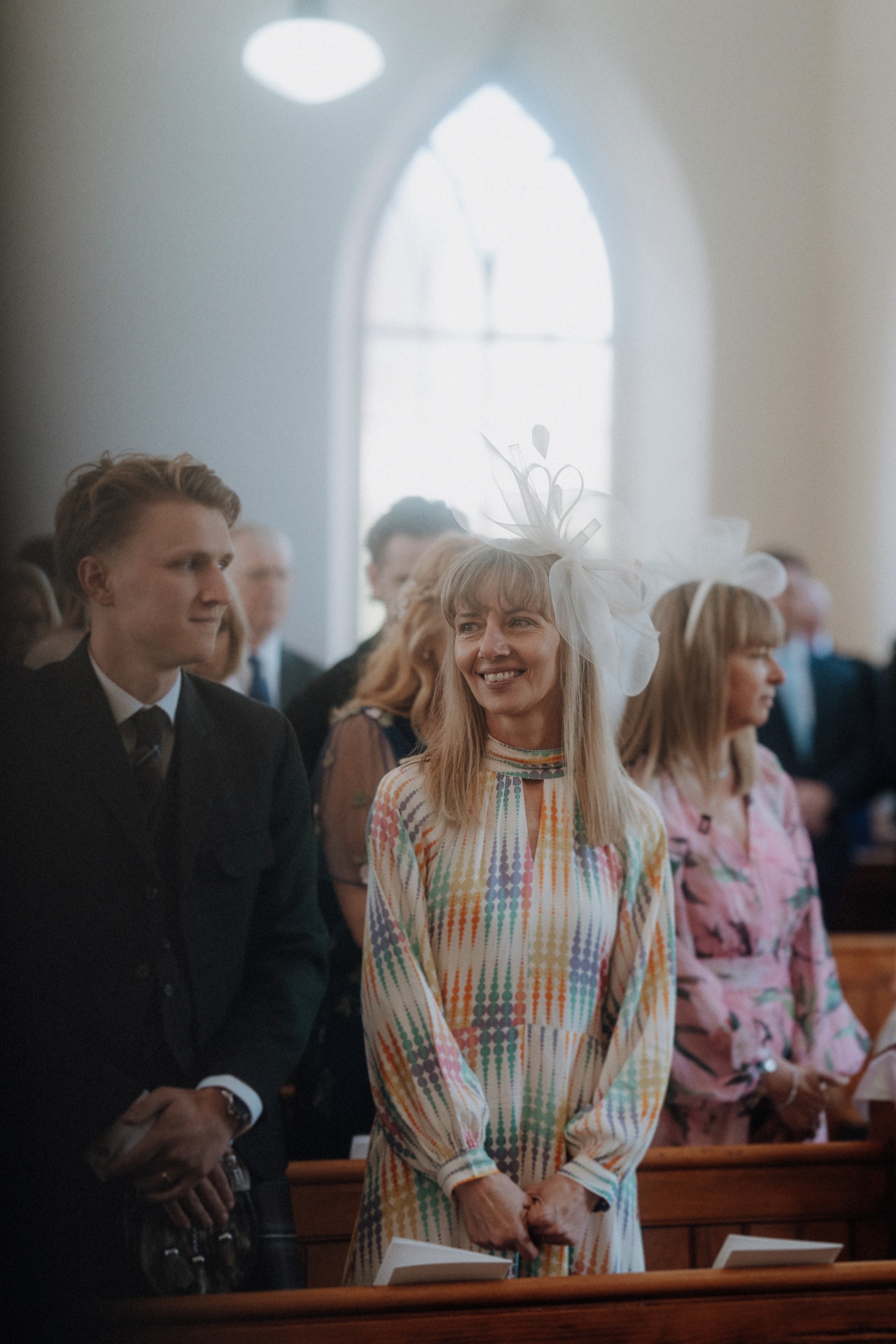 People dressed formally are standing inside a church, with a woman in a patterned dress and white fascinator smiling in the foreground. Light streams through an arched window behind them.