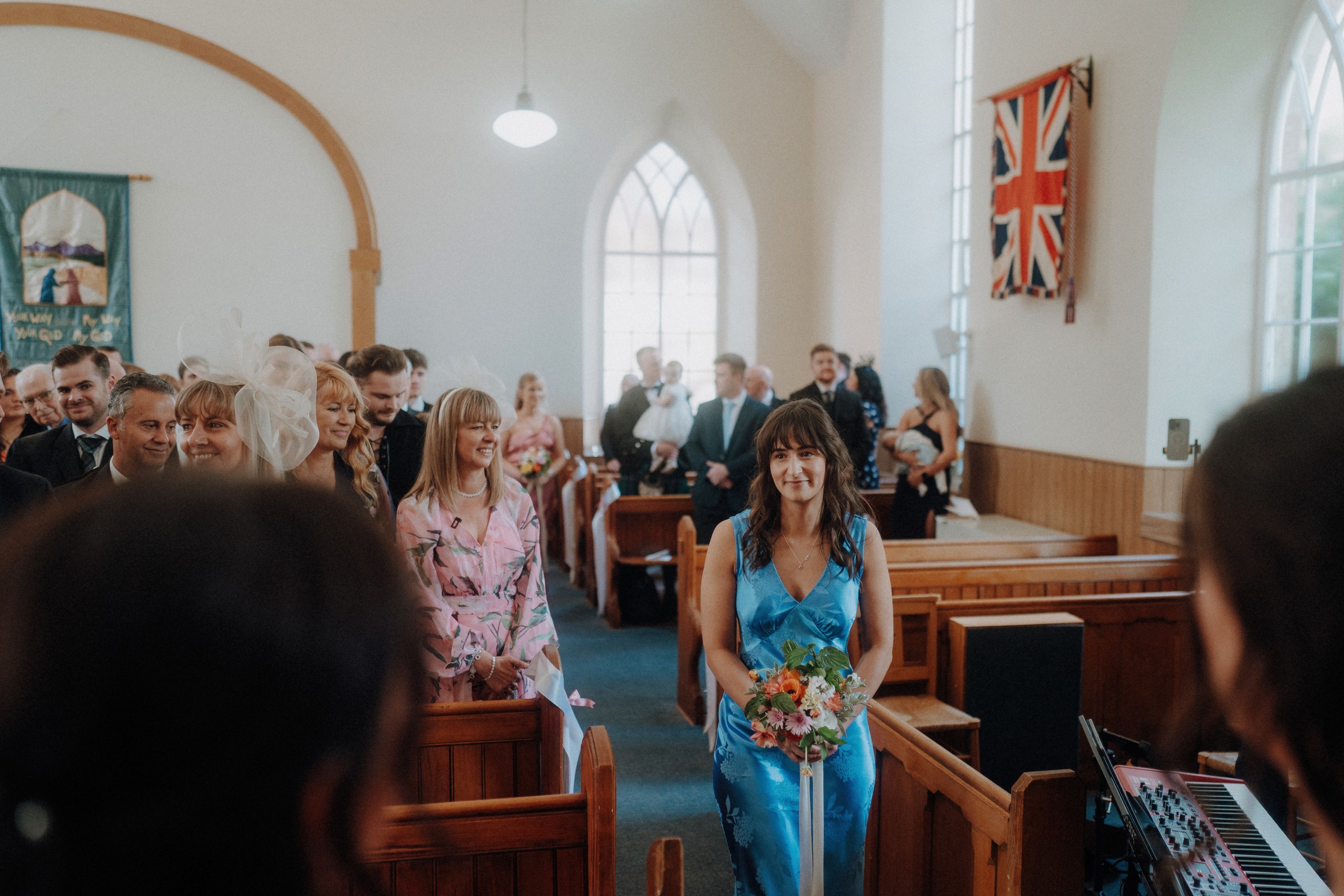 A woman in a blue dress walks down the aisle holding a bouquet in a church filled with seated and standing guests, with a large Union Jack flag on the wall.
