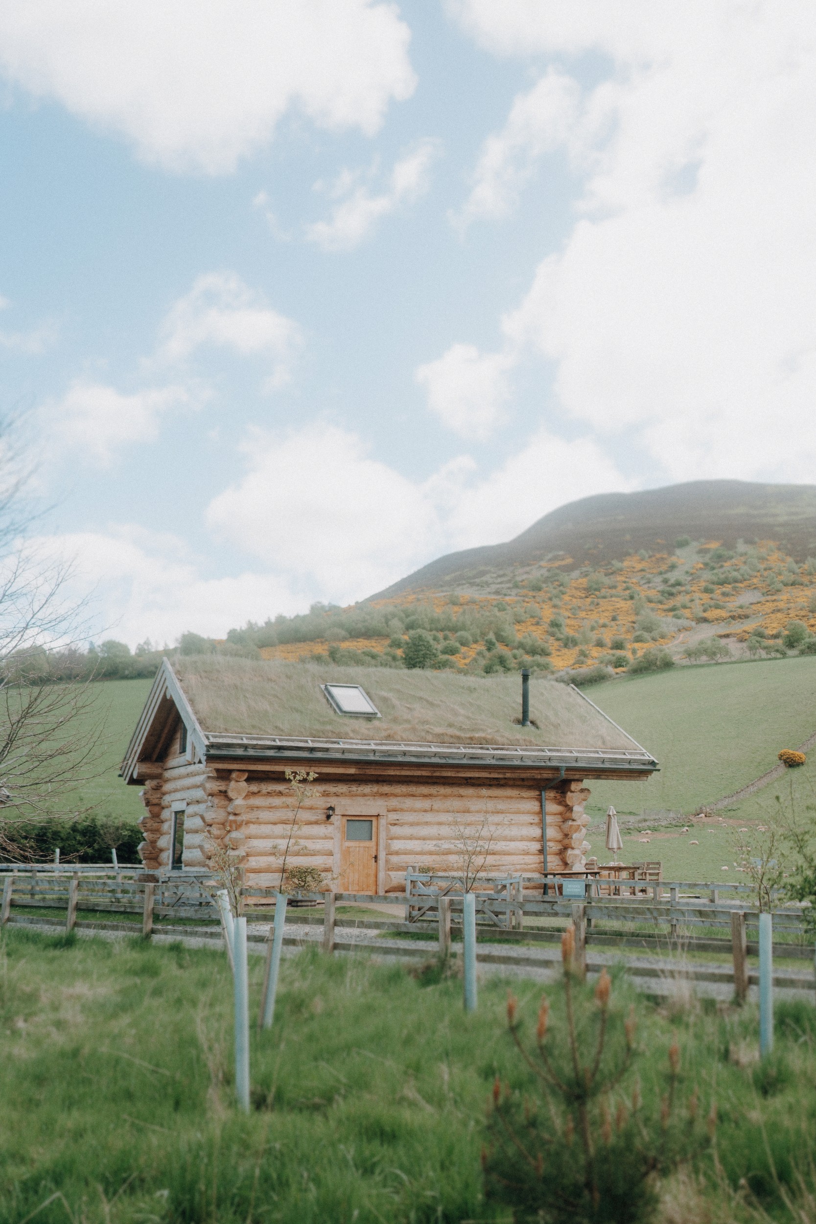 A wooden log cabin with a grass roof sits in a green field near a hillside under a partly cloudy sky.
