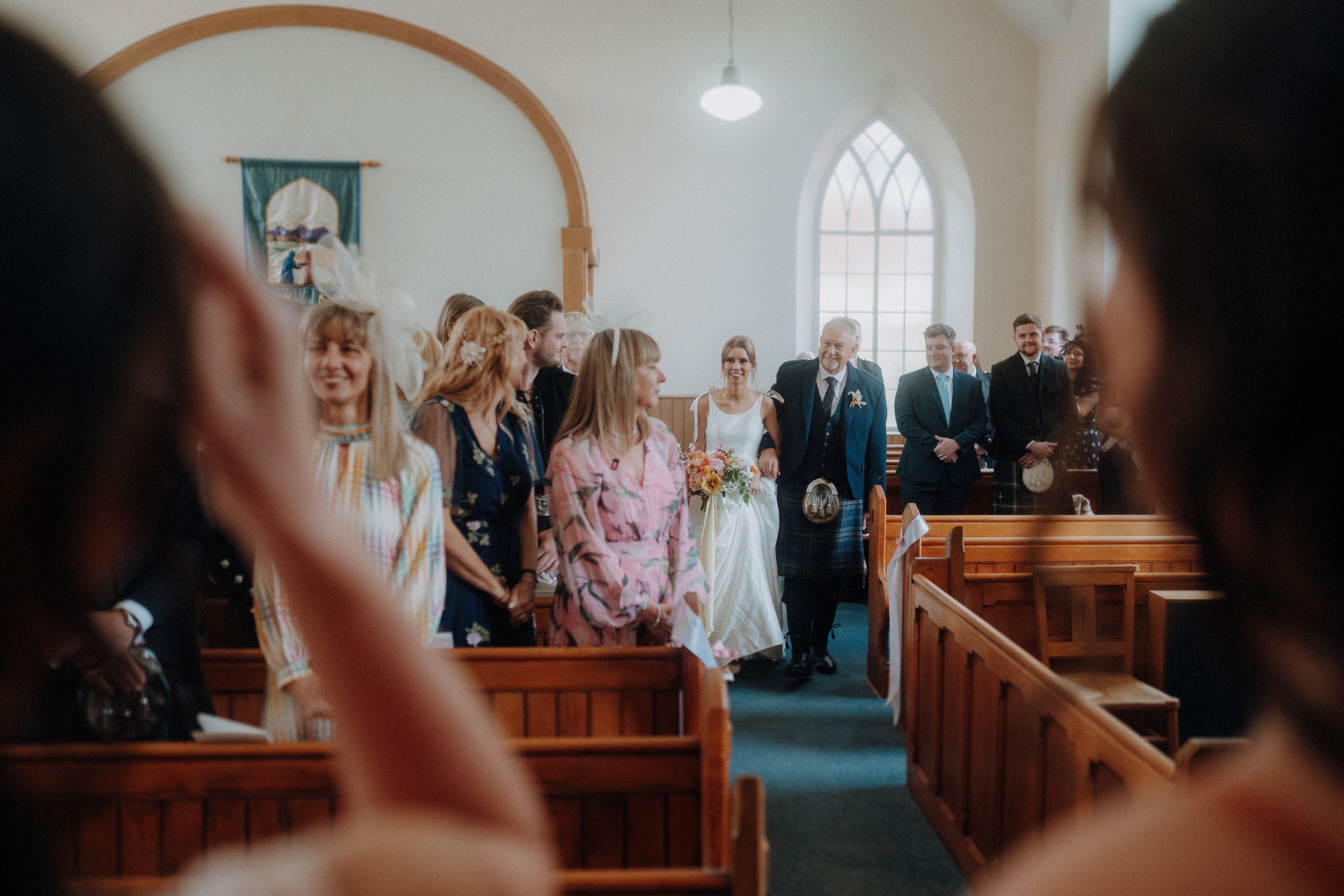 A bride holding a bouquet walks down the aisle with an older man in a church as guests look on and take photos.