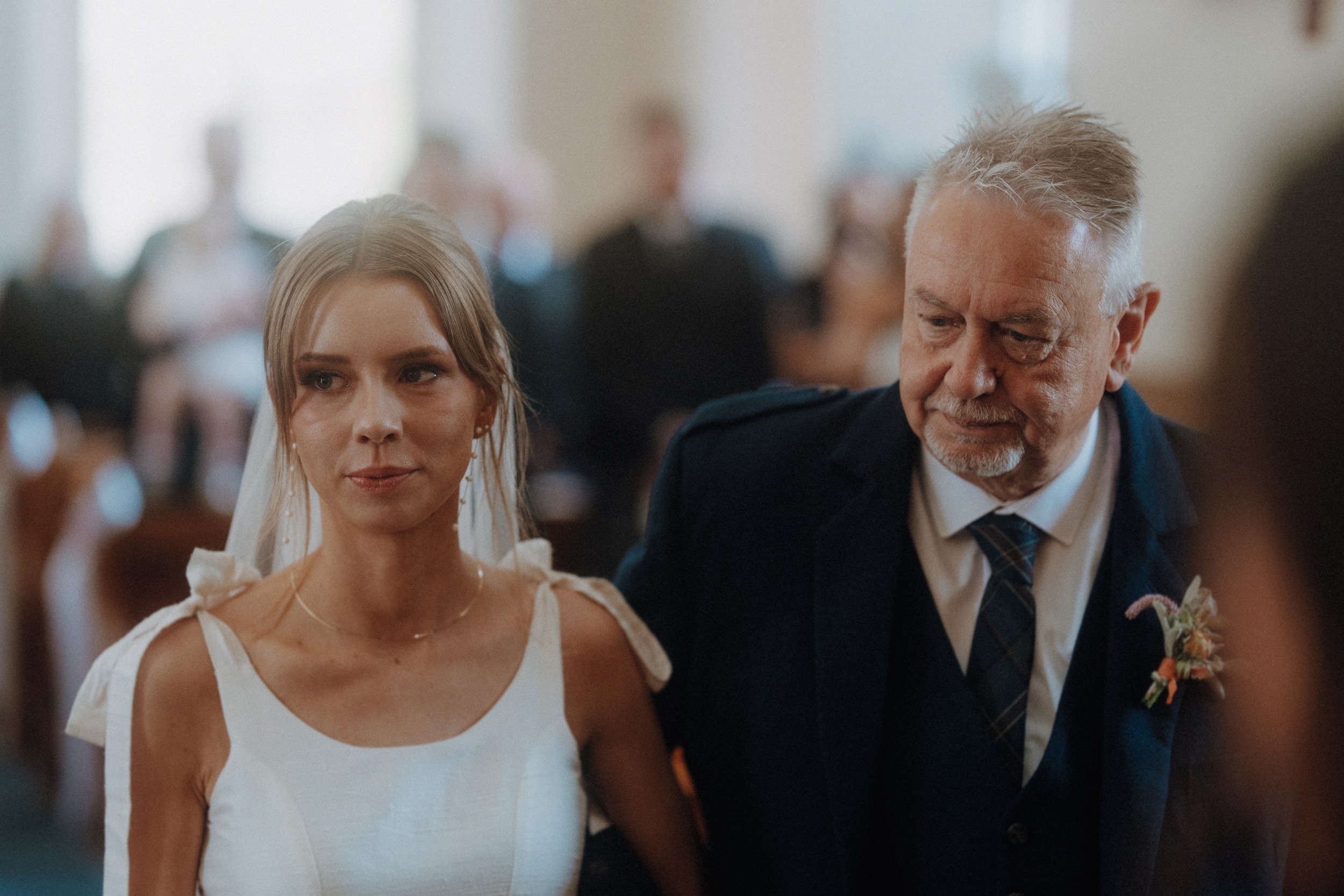 A bride in a white dress stands next to an older man in a suit at a wedding ceremony, with guests blurred in the background.