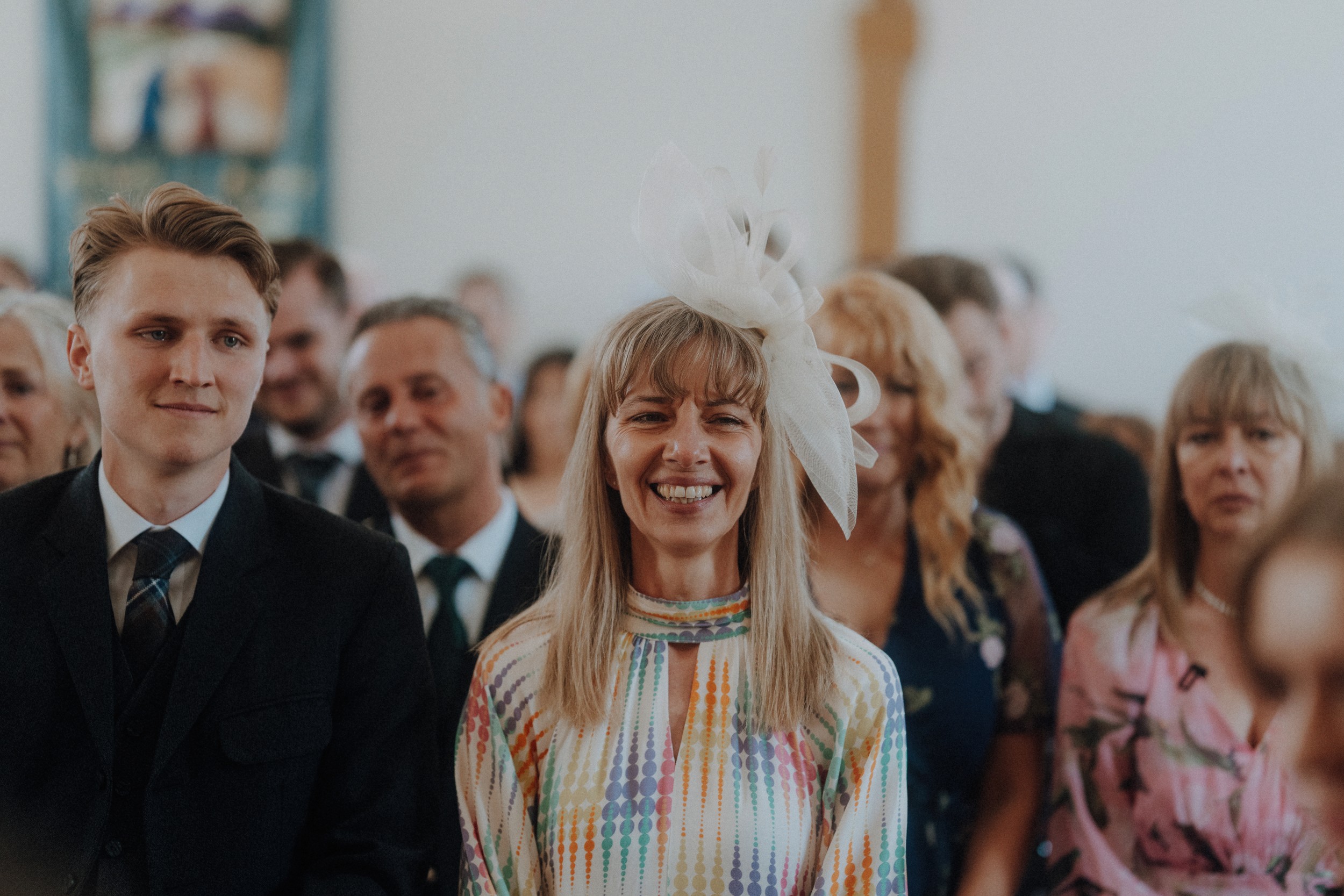 A group of people dressed formally sit and smile at an indoor event; a woman in the center wears a colorful dress and a large white fascinator.