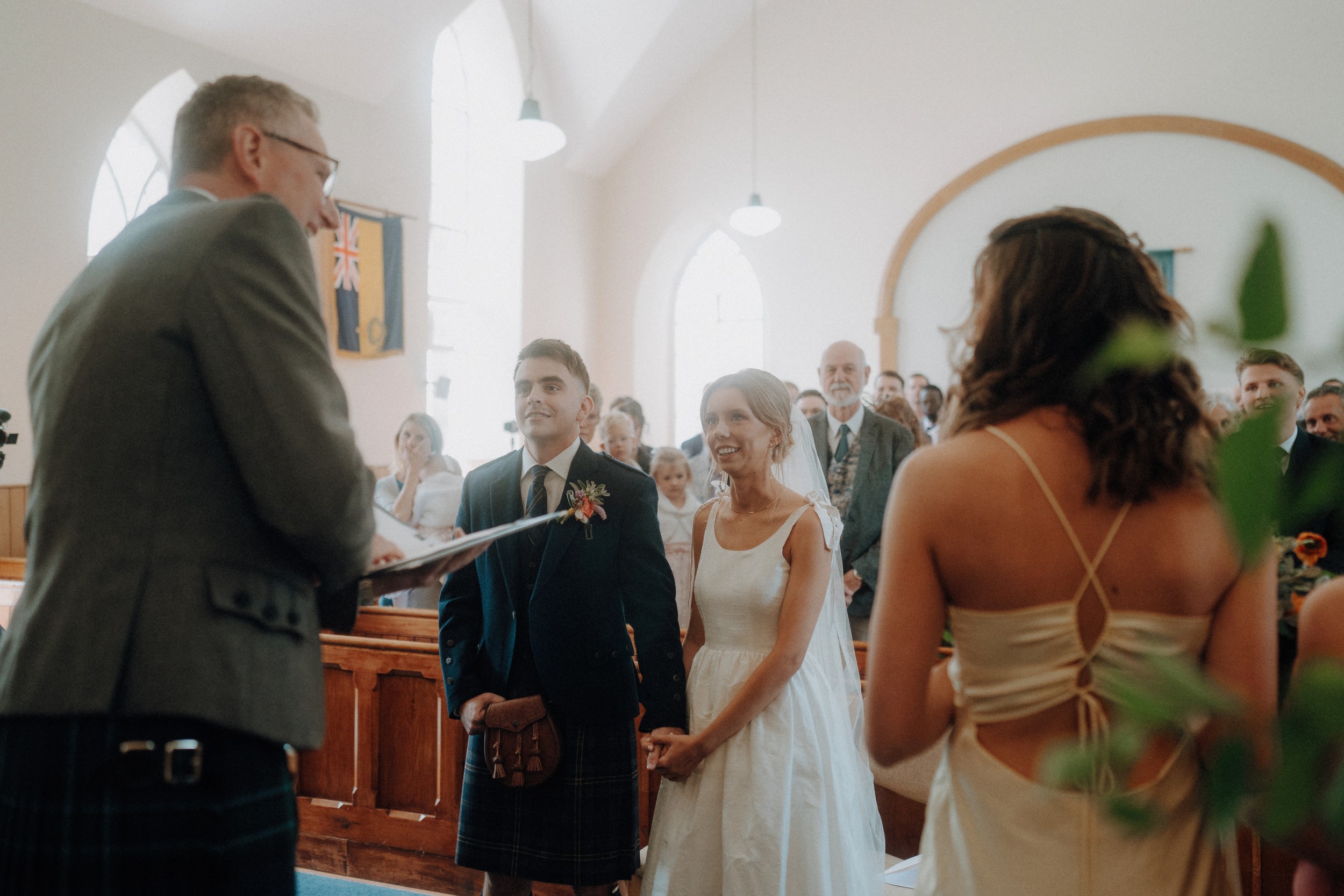 A couple stands holding hands at the altar during a wedding ceremony in a sunlit church, with a celebrant reading and guests seated in the background.