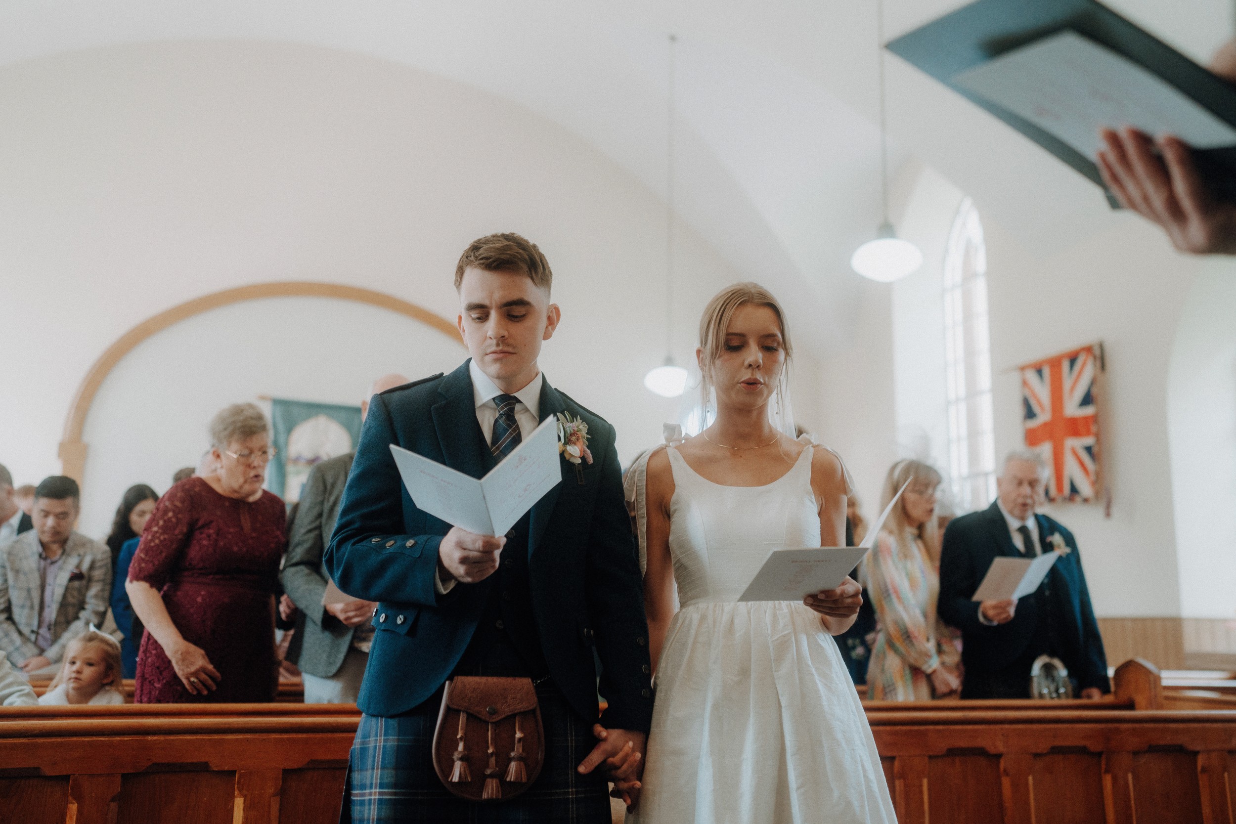 A couple stands at the front of a church holding papers, surrounded by seated and standing guests during a ceremony.