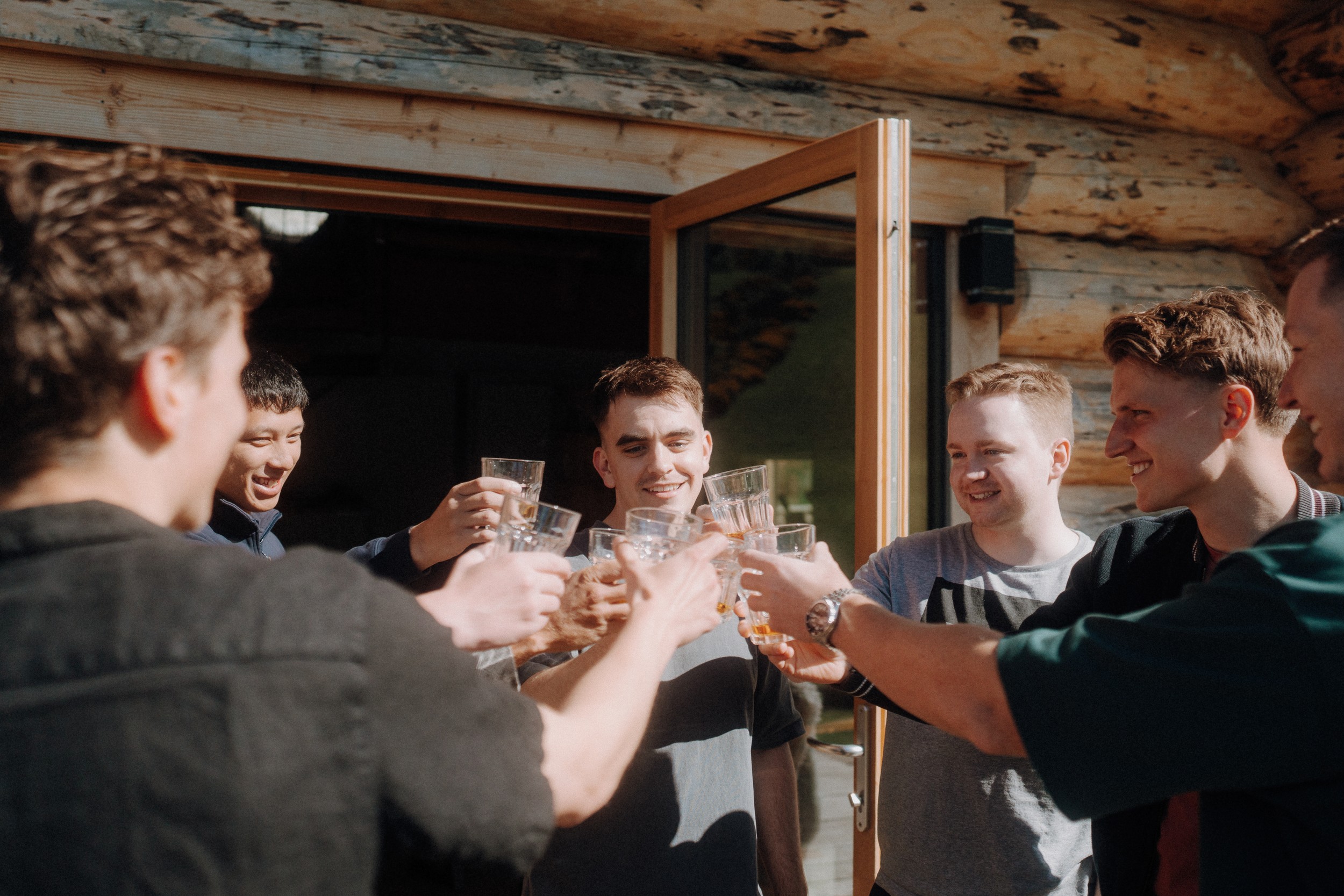 A group of young men stand outdoors in a circle, smiling and raising glasses for a toast near a wooden building on a sunny day.