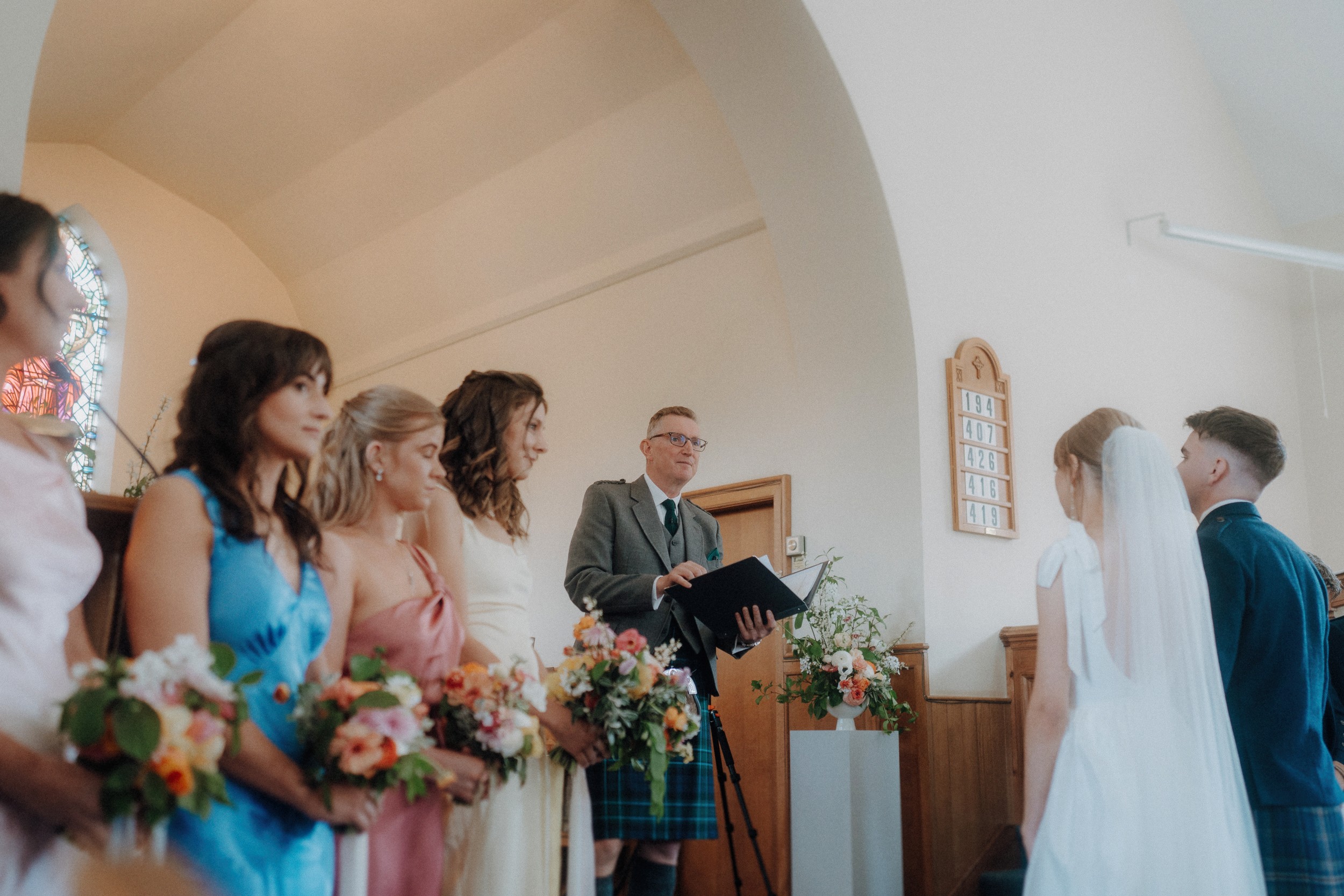 A wedding ceremony in a church, with bridesmaids in colorful dresses holding bouquets and a man officiating as the bride and groom stand together.