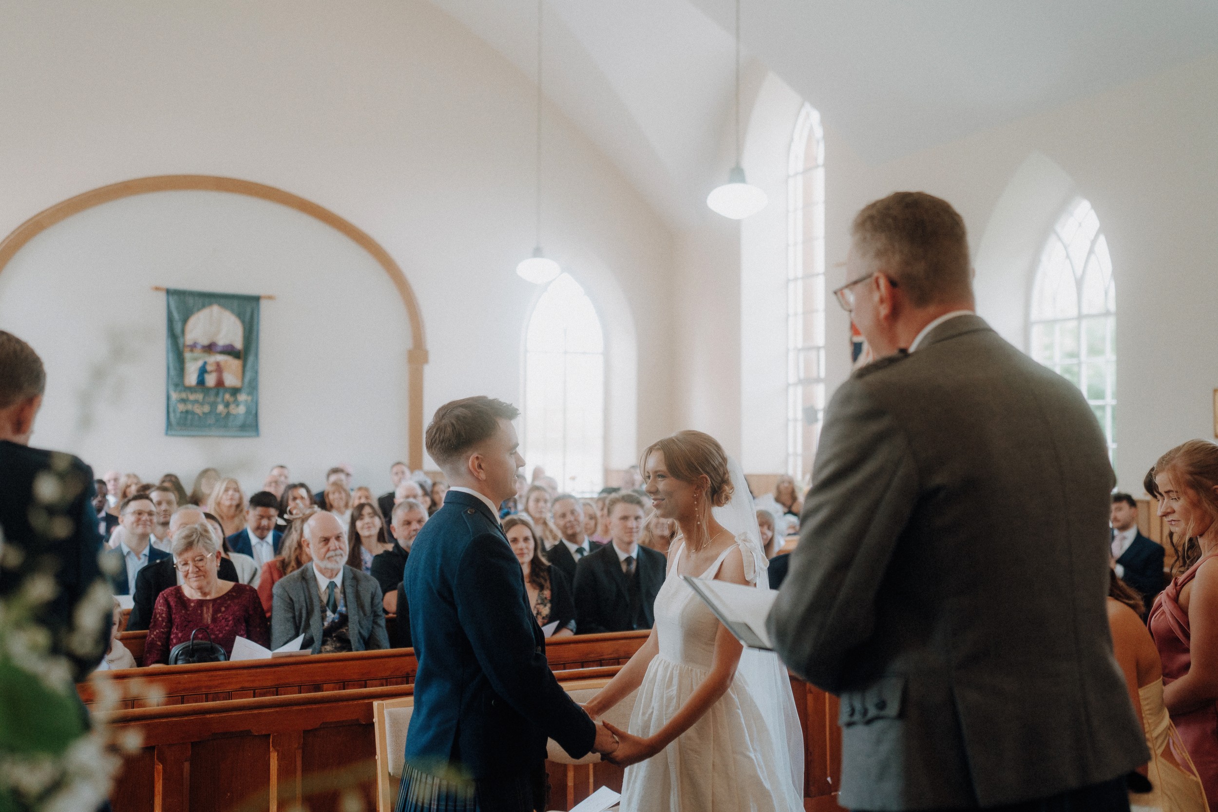 A couple stands facing each other and holding hands at the altar during a wedding ceremony in a church, with guests seated and watching.