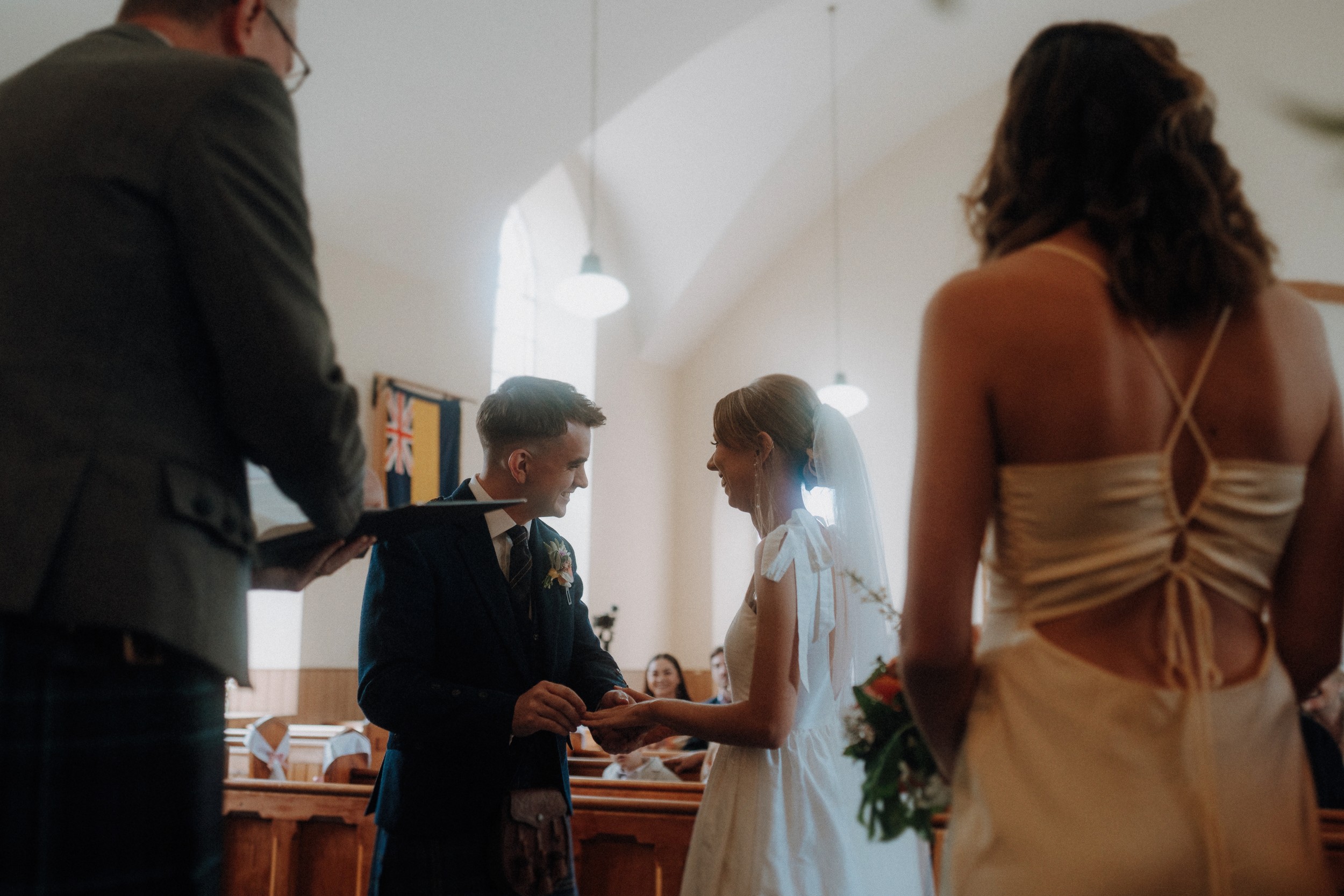 A couple stands at the altar exchanging rings during a wedding ceremony, with a person officiating and another guest standing nearby.
