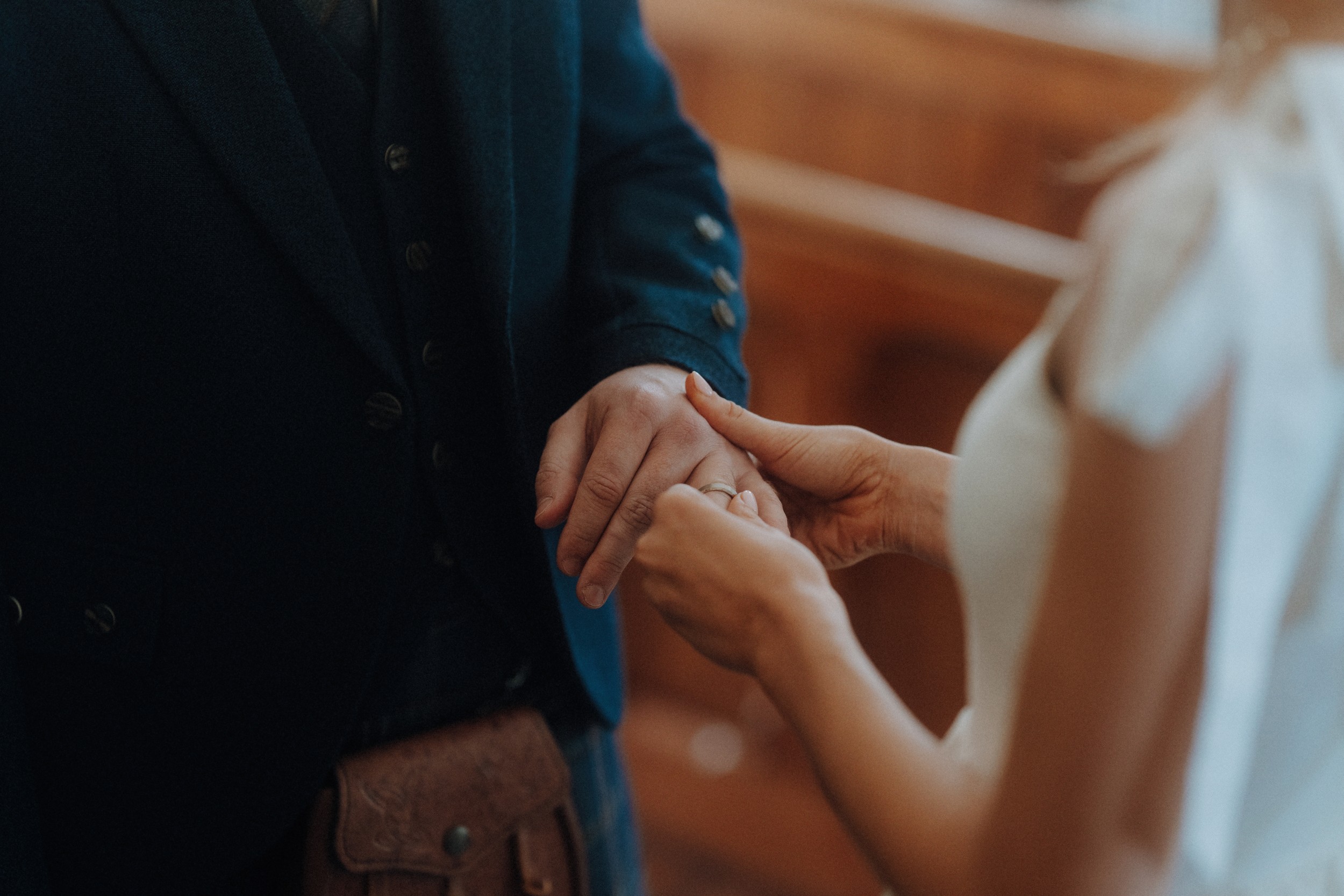 A person in a suit is having a ring placed on their finger by another person in a white dress, suggesting a wedding ceremony.