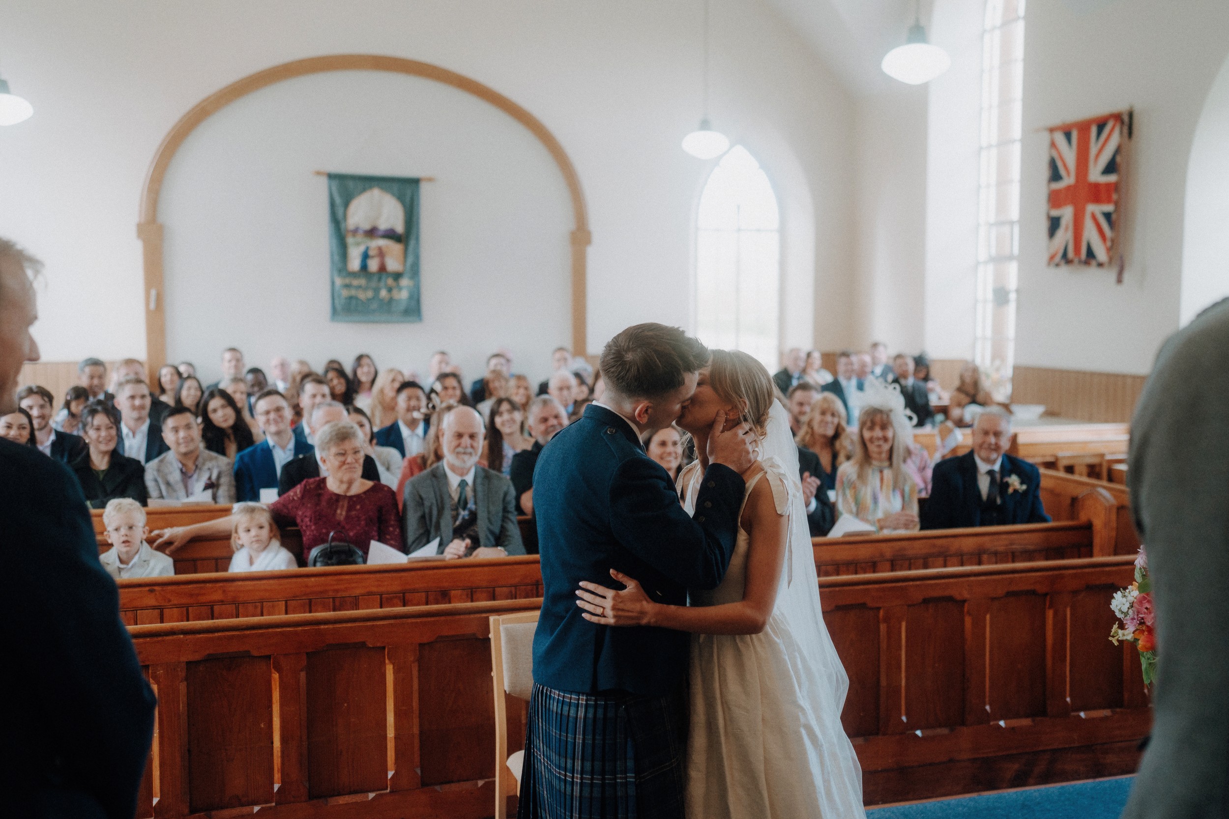 A bride and groom kiss at the altar in a church filled with seated guests, with a large window and a Union Jack flag visible in the background.