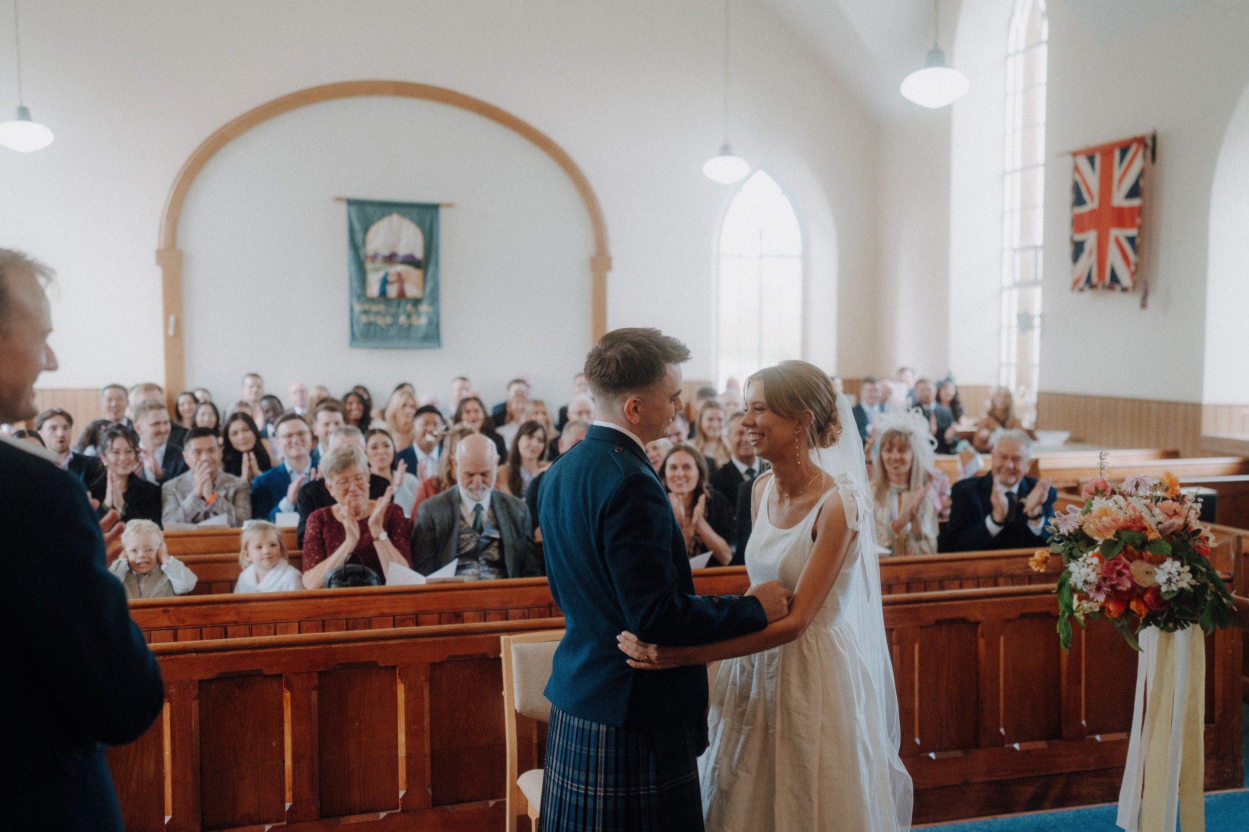 A bride and groom stand facing each other at the front of a church, surrounded by seated guests who are clapping.