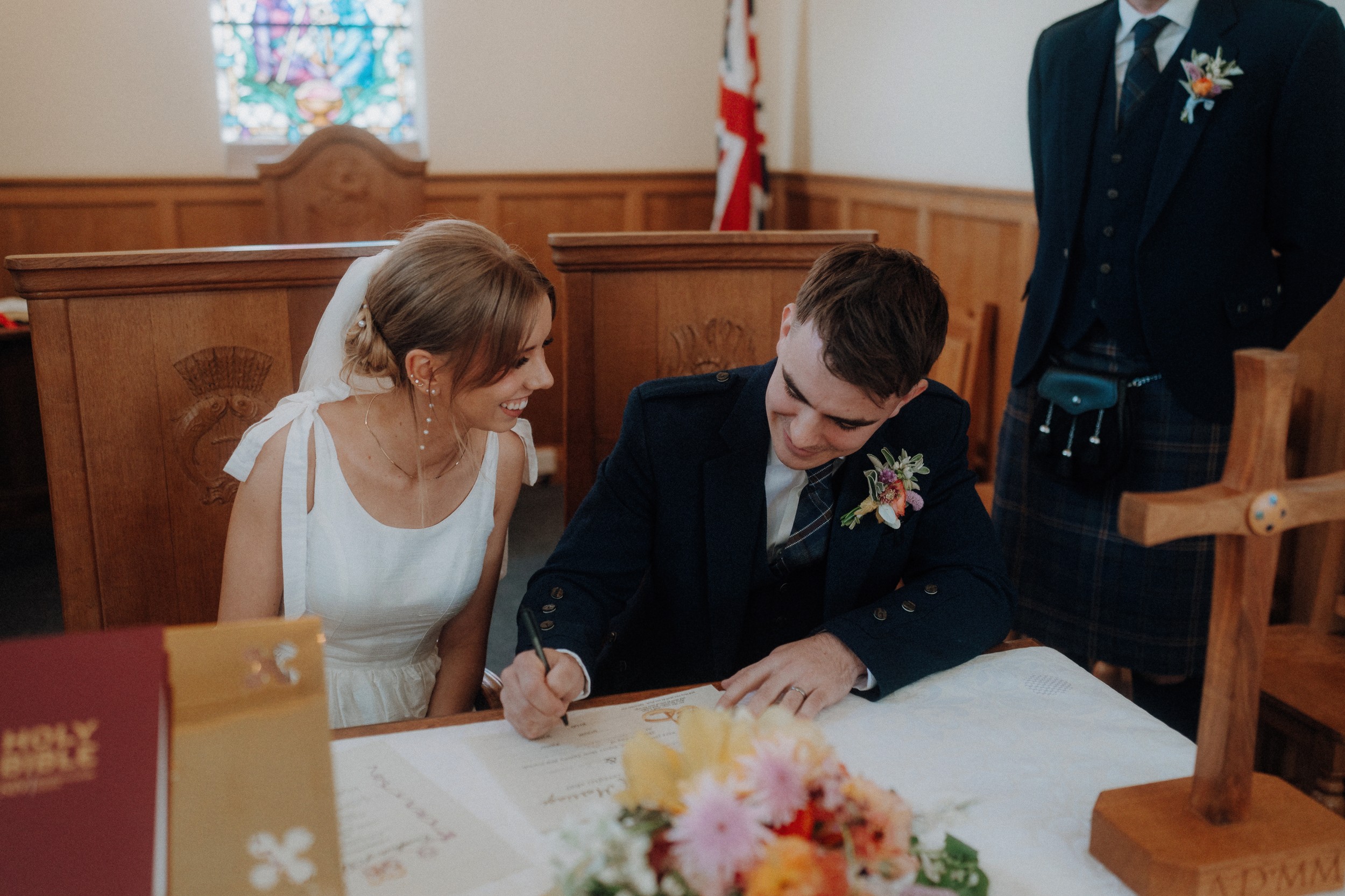 A bride and groom sit at a table in a church while the groom signs a document; the bride smiles and looks at him. A man in a kilt stands nearby.