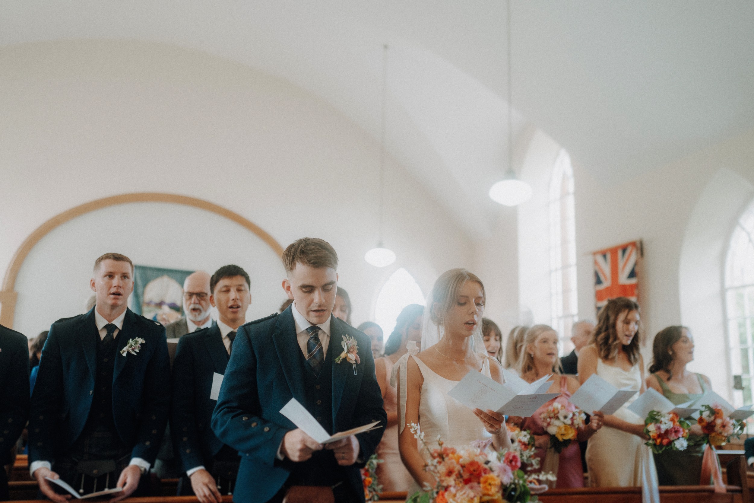 A group of people dressed formally stand in church pews, holding papers and singing; floral arrangements and flags are visible in the background.