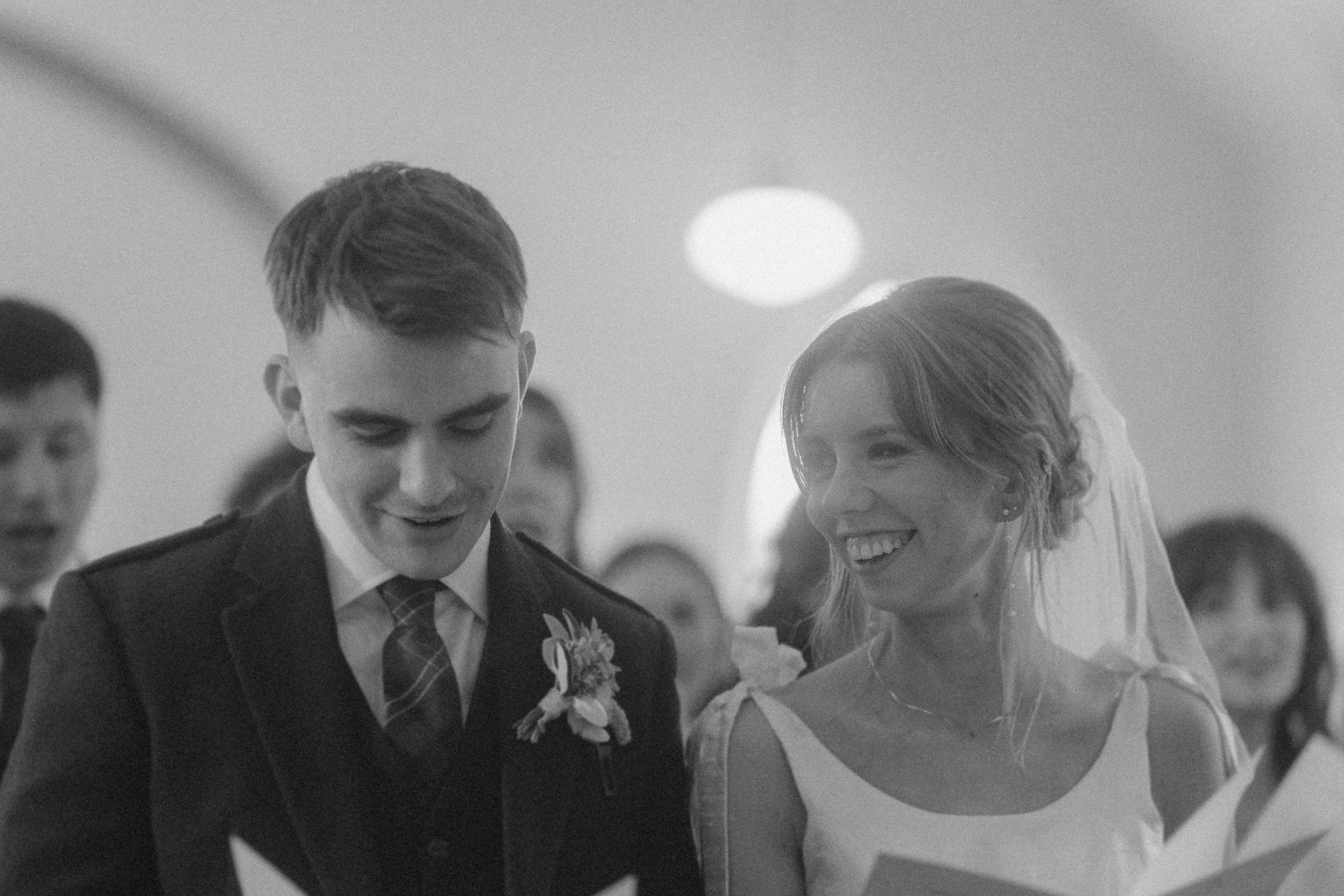 A bride and groom stand together at their wedding ceremony, smiling and holding papers, with other people visible in the background.