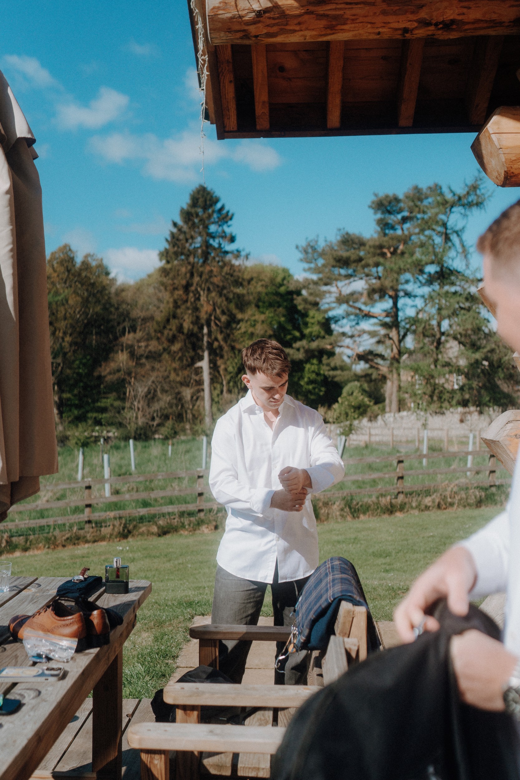 A man outdoors in a white dress shirt adjusts his sleeves near a wooden building, with another person in the foreground and clothing items on a picnic table.