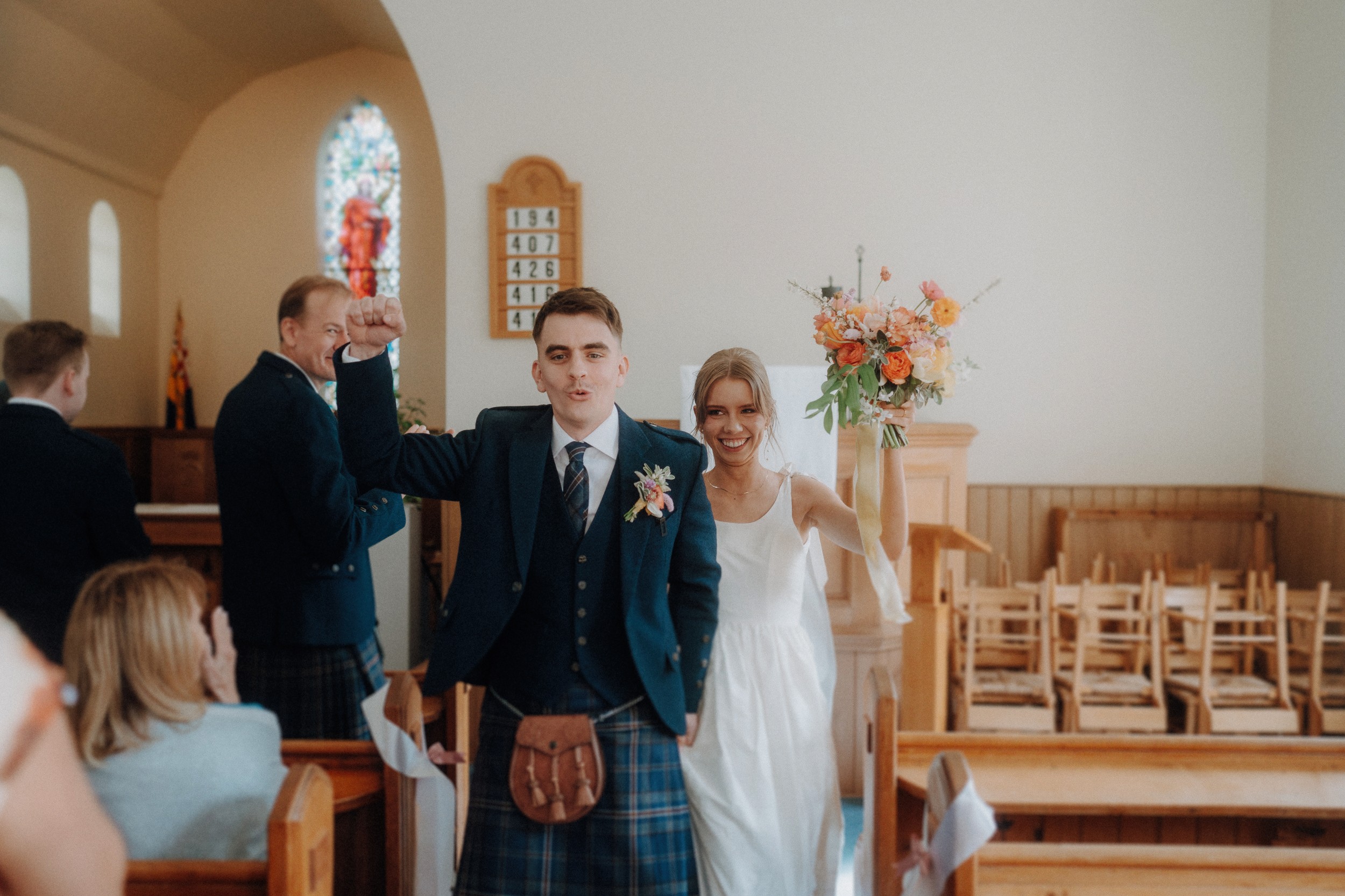 A newly married couple walks down the aisle of a church, the groom raising his fist and the bride holding a bouquet, while guests look on.