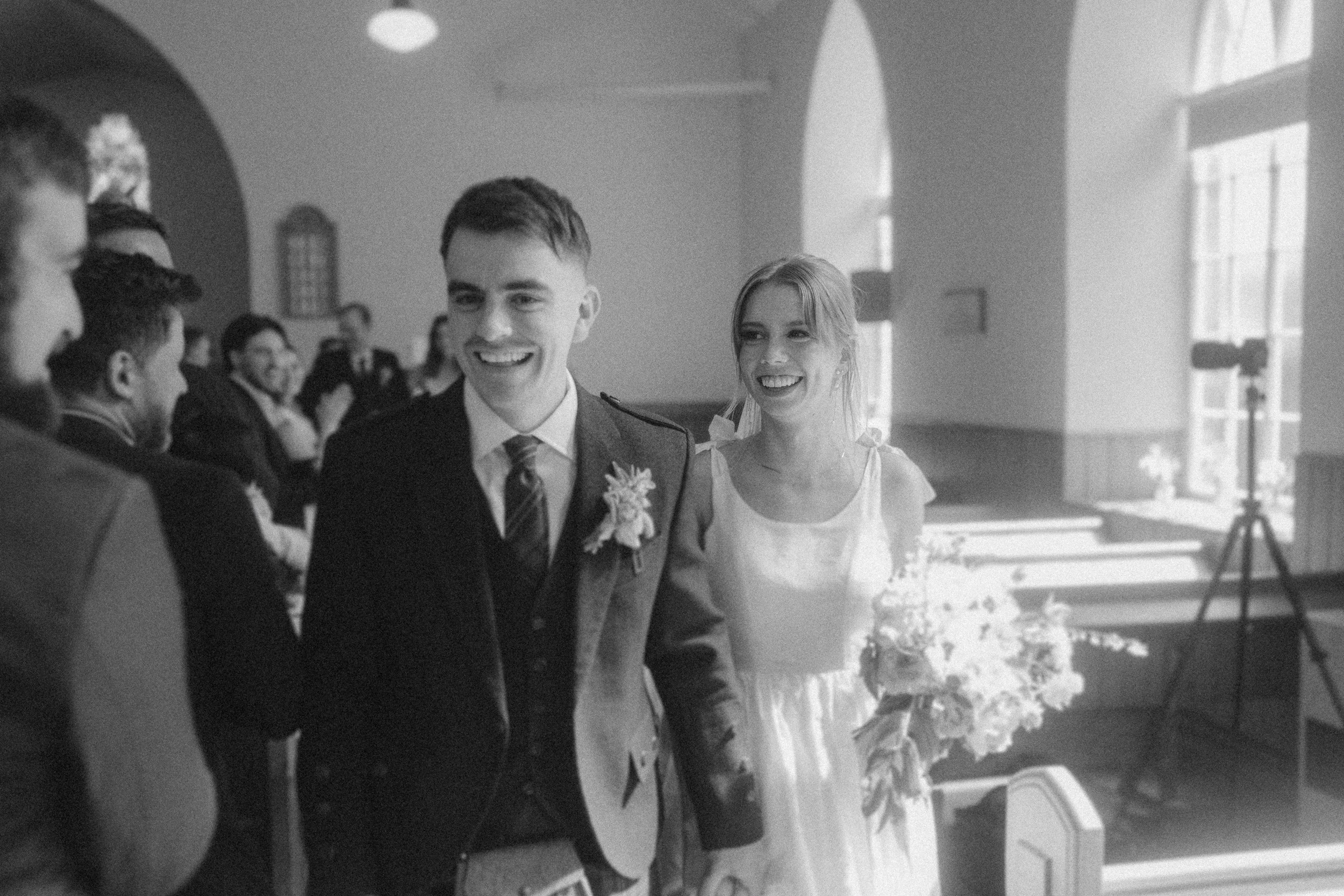 A bride and groom walk down the aisle smiling, holding hands, with guests watching from pews in a sunlit church.