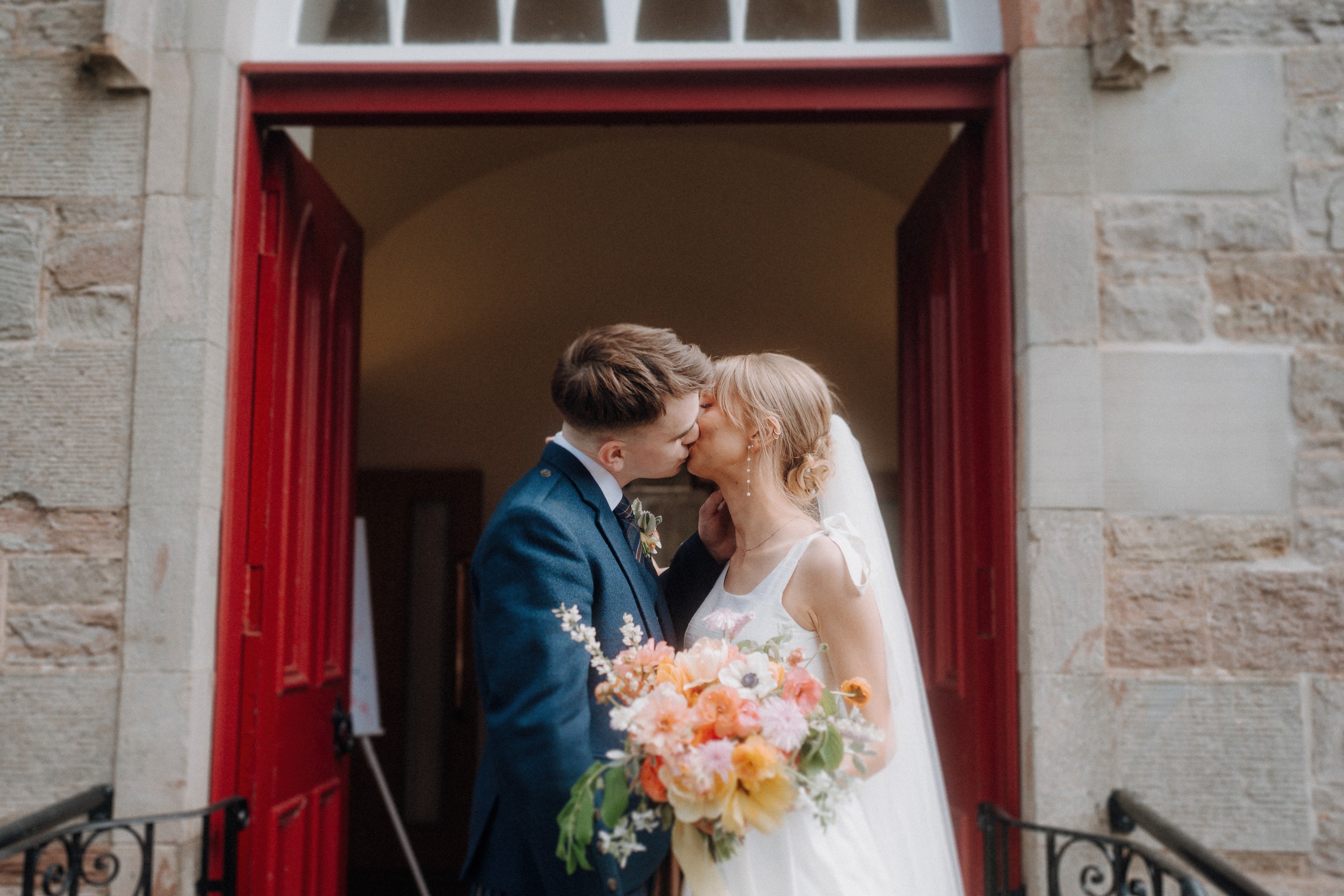 A bride and groom kiss while standing in front of open red doors, with the bride holding a bouquet of flowers.