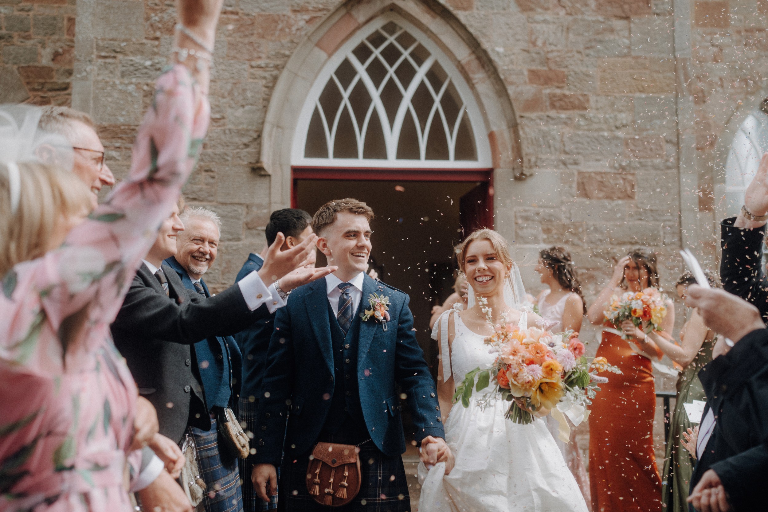 A newlywed couple walks out of a stone church as guests celebrate and throw confetti. The bride holds a bouquet and both are smiling.