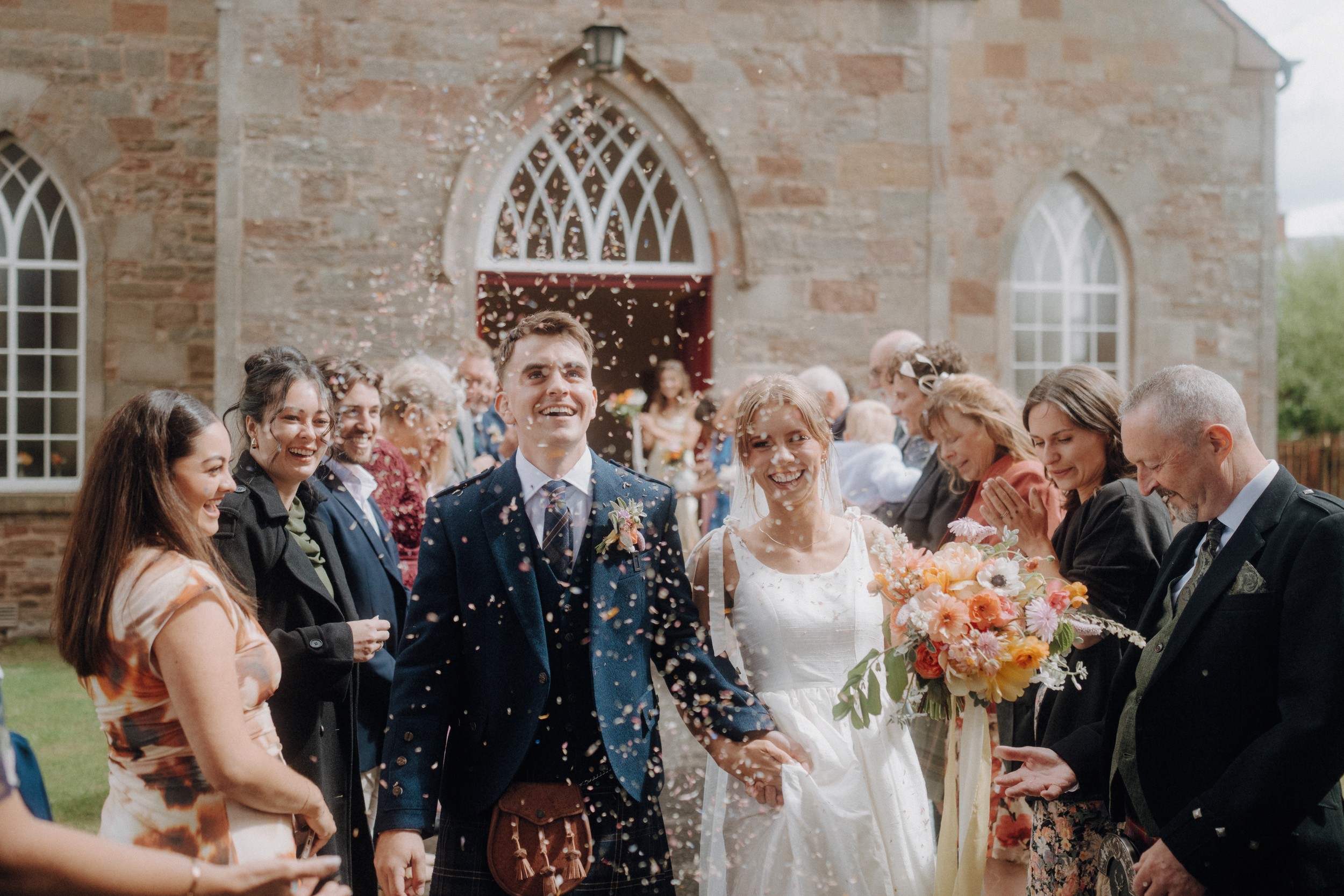 A bride and groom walk outside a stone church as guests throw confetti and smile, with the bride holding a colorful bouquet.