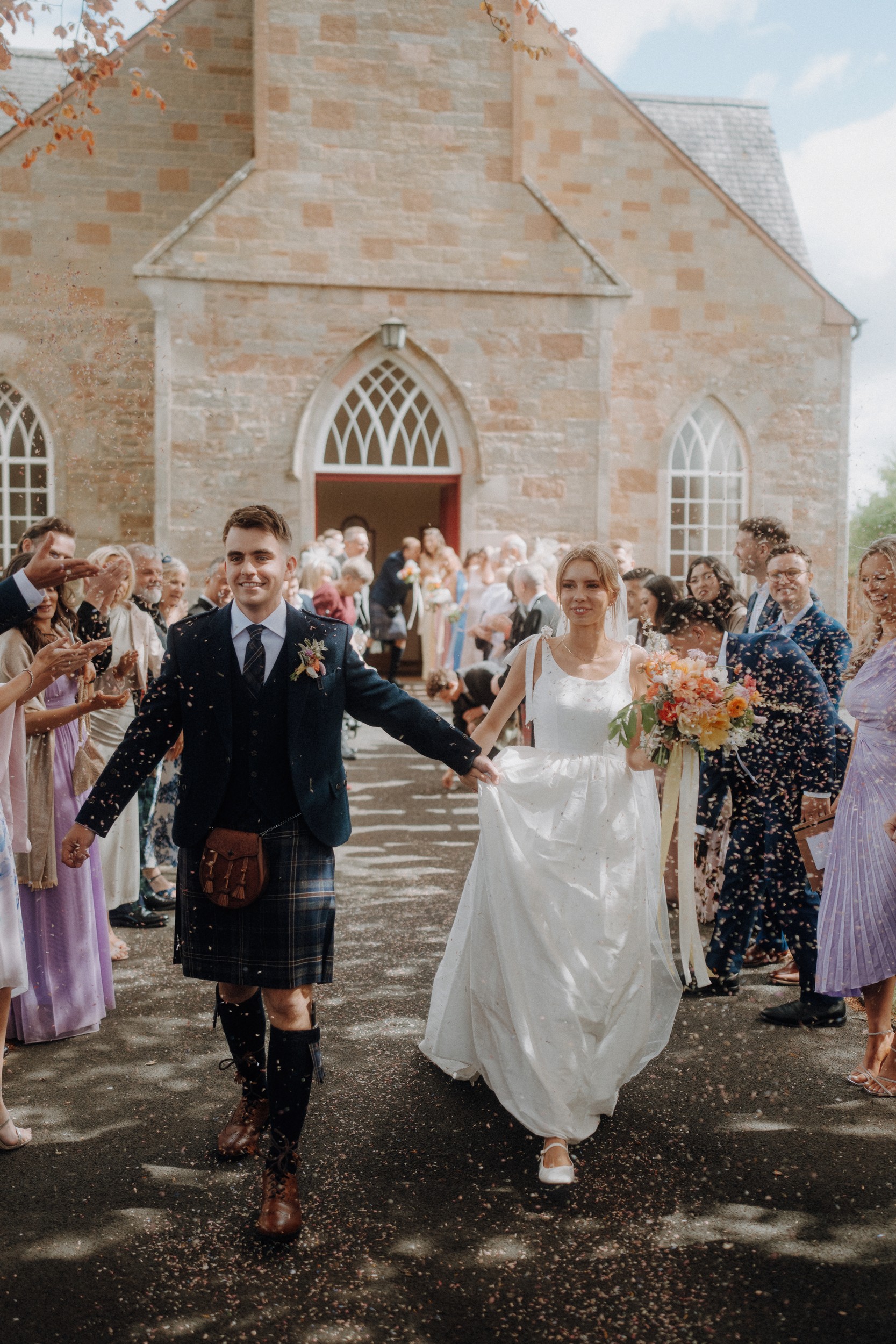 A bride and groom walk hand in hand outside a stone church, surrounded by guests throwing confetti. The groom wears a kilt and the bride holds a colorful bouquet.