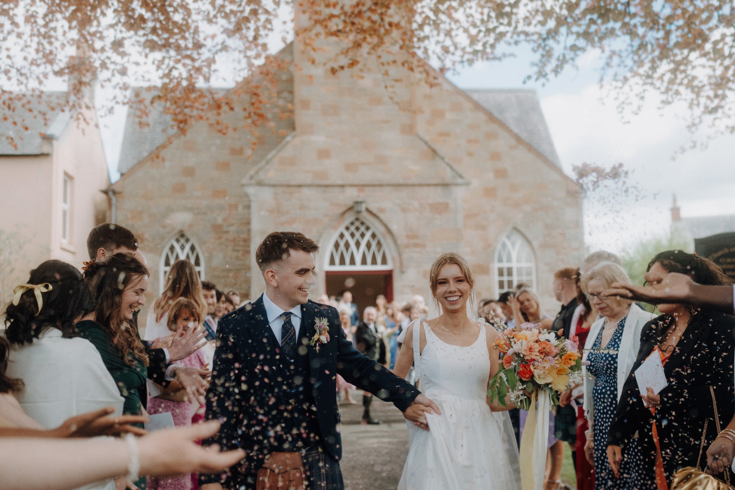 Bride and groom walk hand in hand outside a church while guests throw confetti and celebrate around them.