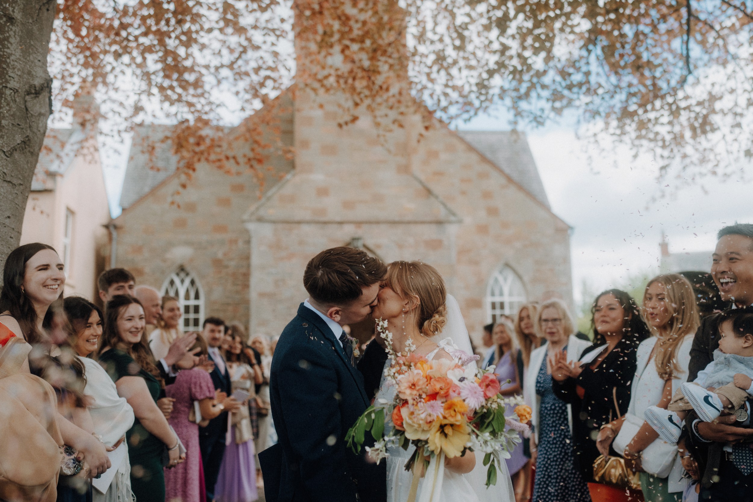 A bride and groom kiss outdoors in front of a stone building, holding a bouquet, surrounded by smiling guests, some of whom are throwing confetti.
