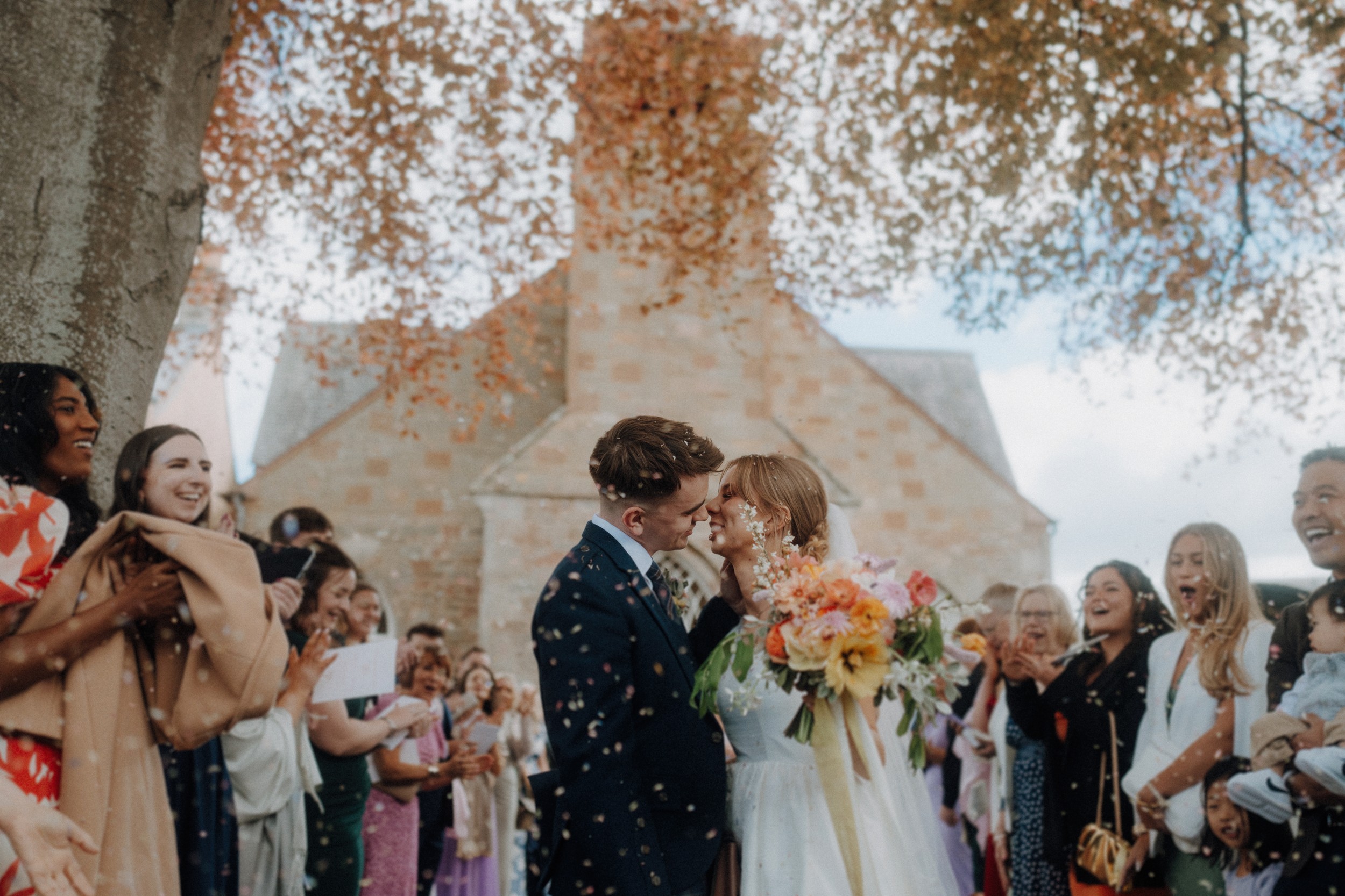 A bride and groom kiss under a tree outside a stone building as guests cheer and throw confetti around them.