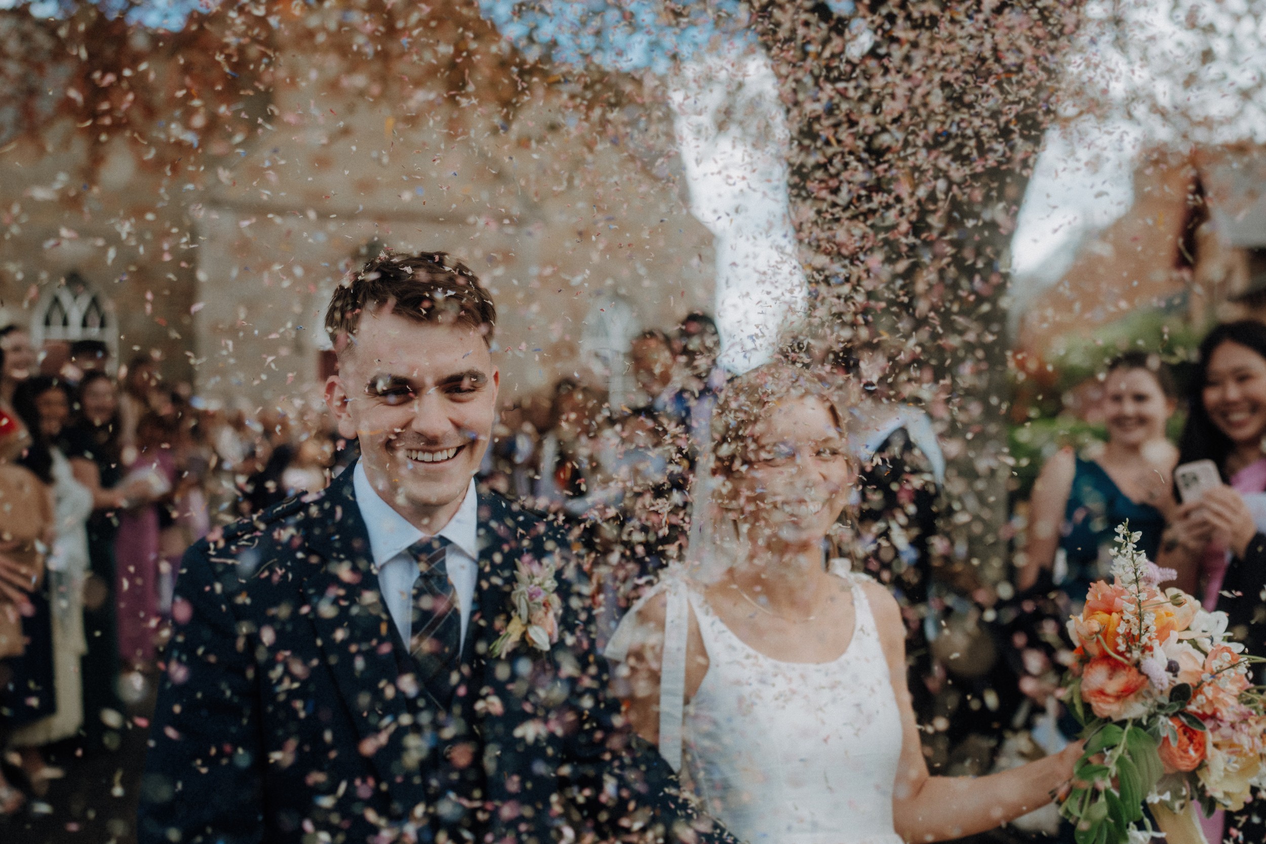A bride and groom walk outdoors while guests throw confetti. The bride holds a bouquet of flowers, and both are smiling amid a crowd of onlookers.