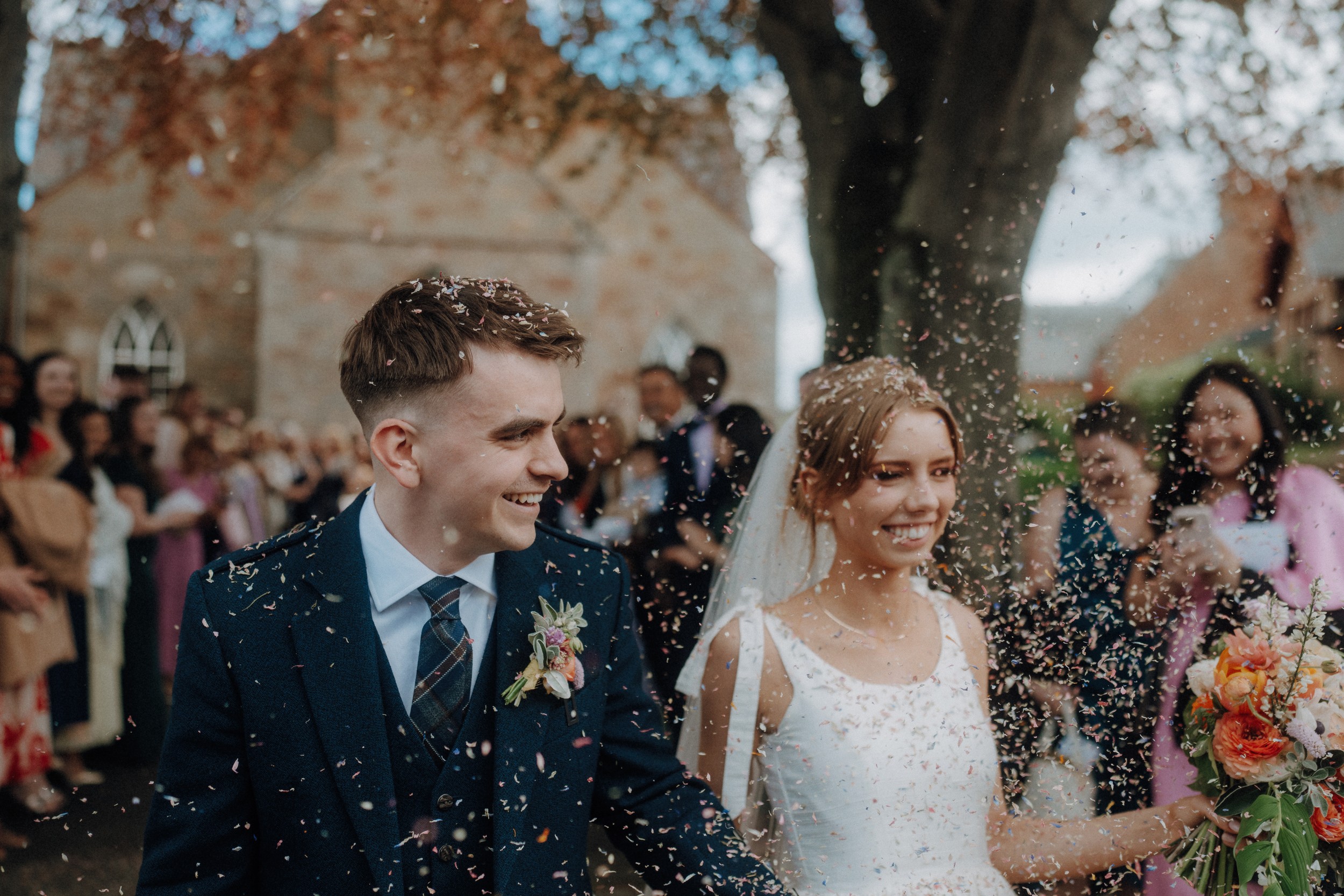 A bride and groom walk outside holding hands, smiling, as confetti is thrown by guests gathered behind them at a wedding ceremony.