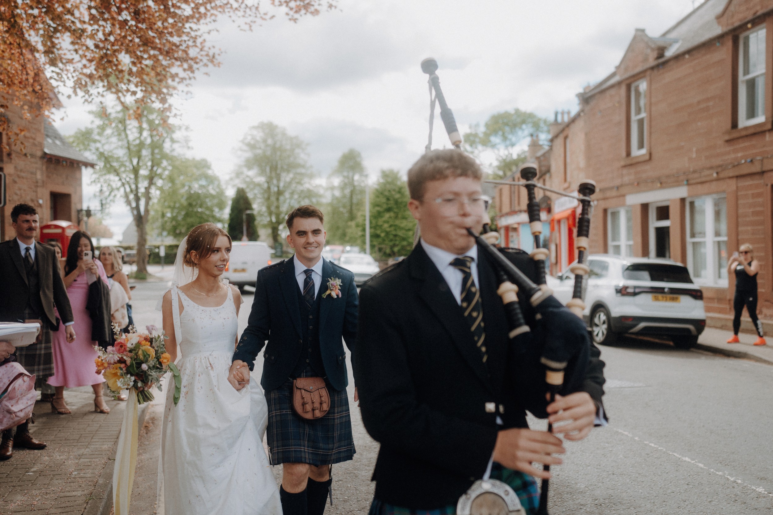 A bride and groom walk hand in hand behind a bagpiper on a sidewalk, followed by guests, with houses and cars visible in the background.