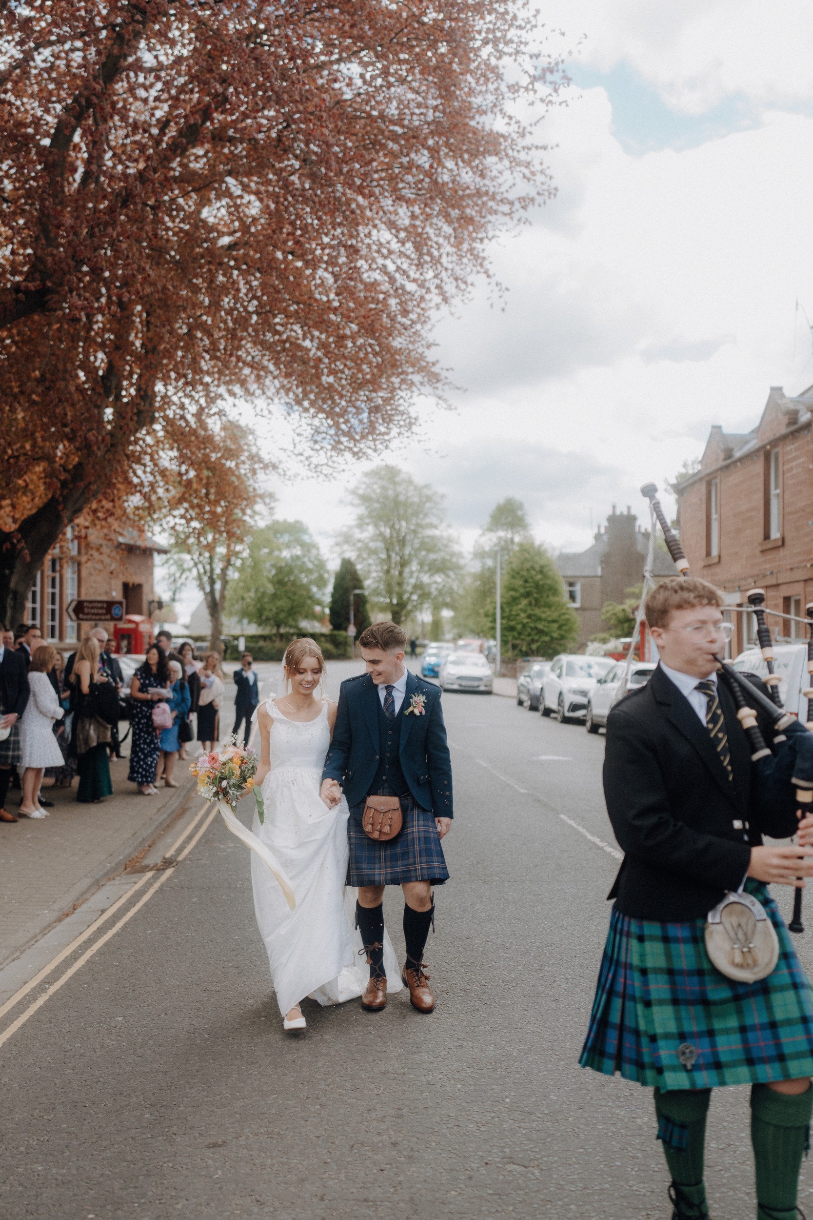 A bride and groom walk down a street, holding hands, followed by guests, while a bagpiper in traditional Scottish attire plays in the foreground.