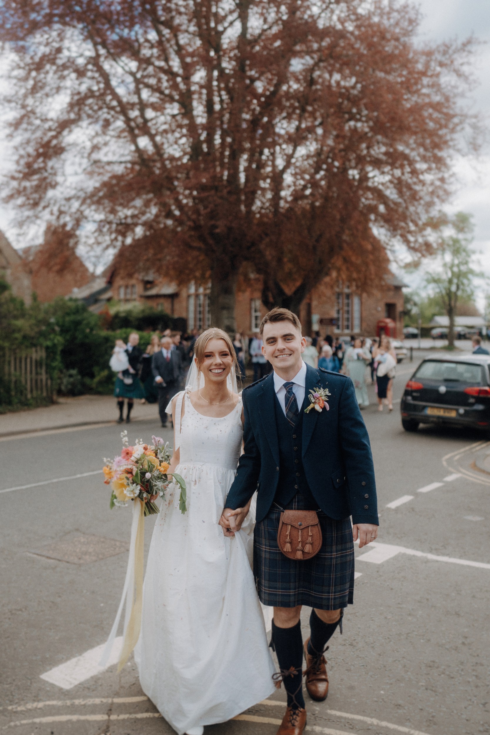 A bride in a white dress and a groom in a kilt walk hand in hand outside, smiling, with guests and parked cars visible in the background.