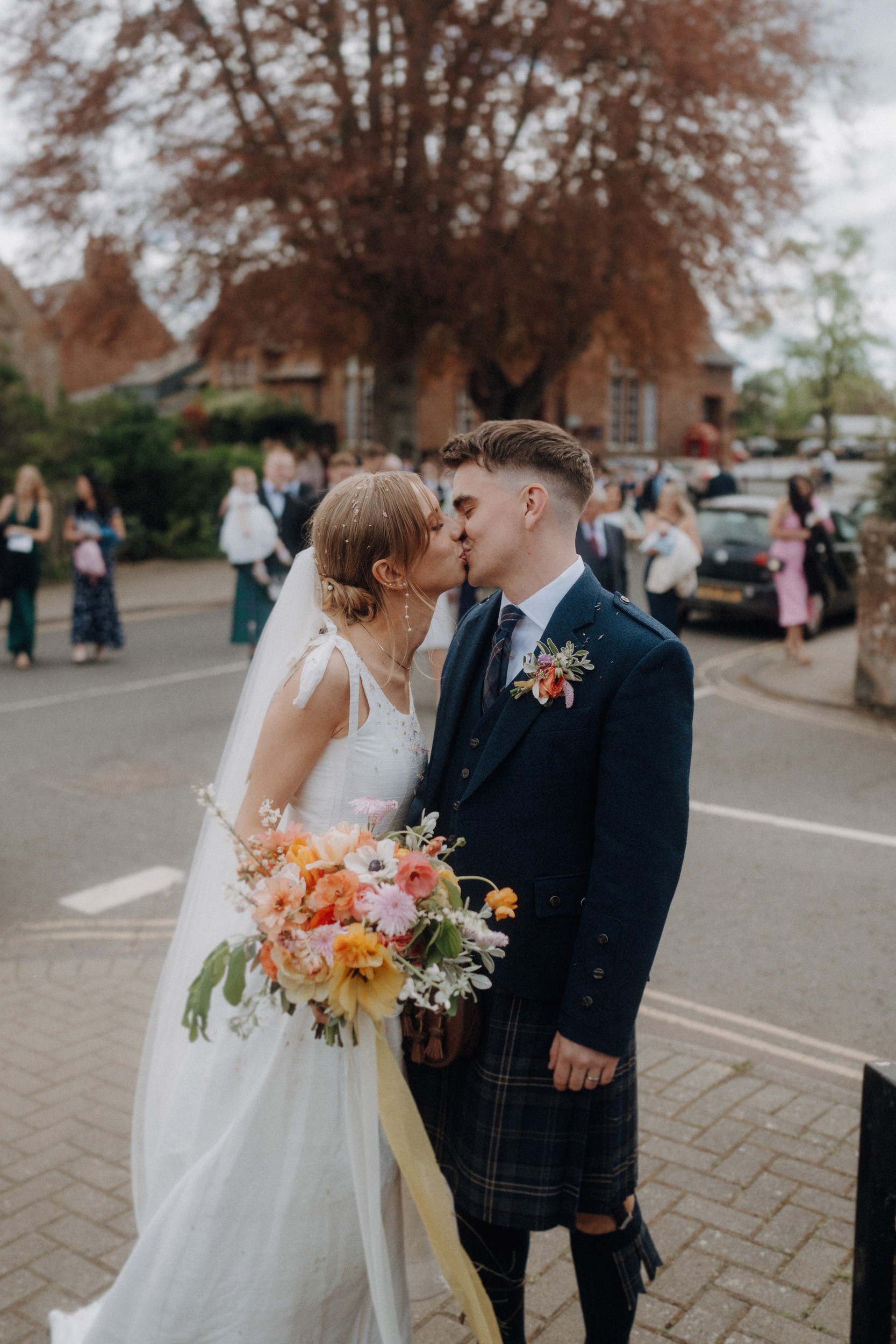 A bride in a white dress and a groom in a kilt share a kiss outdoors, with the bride holding a bouquet and guests visible in the background.