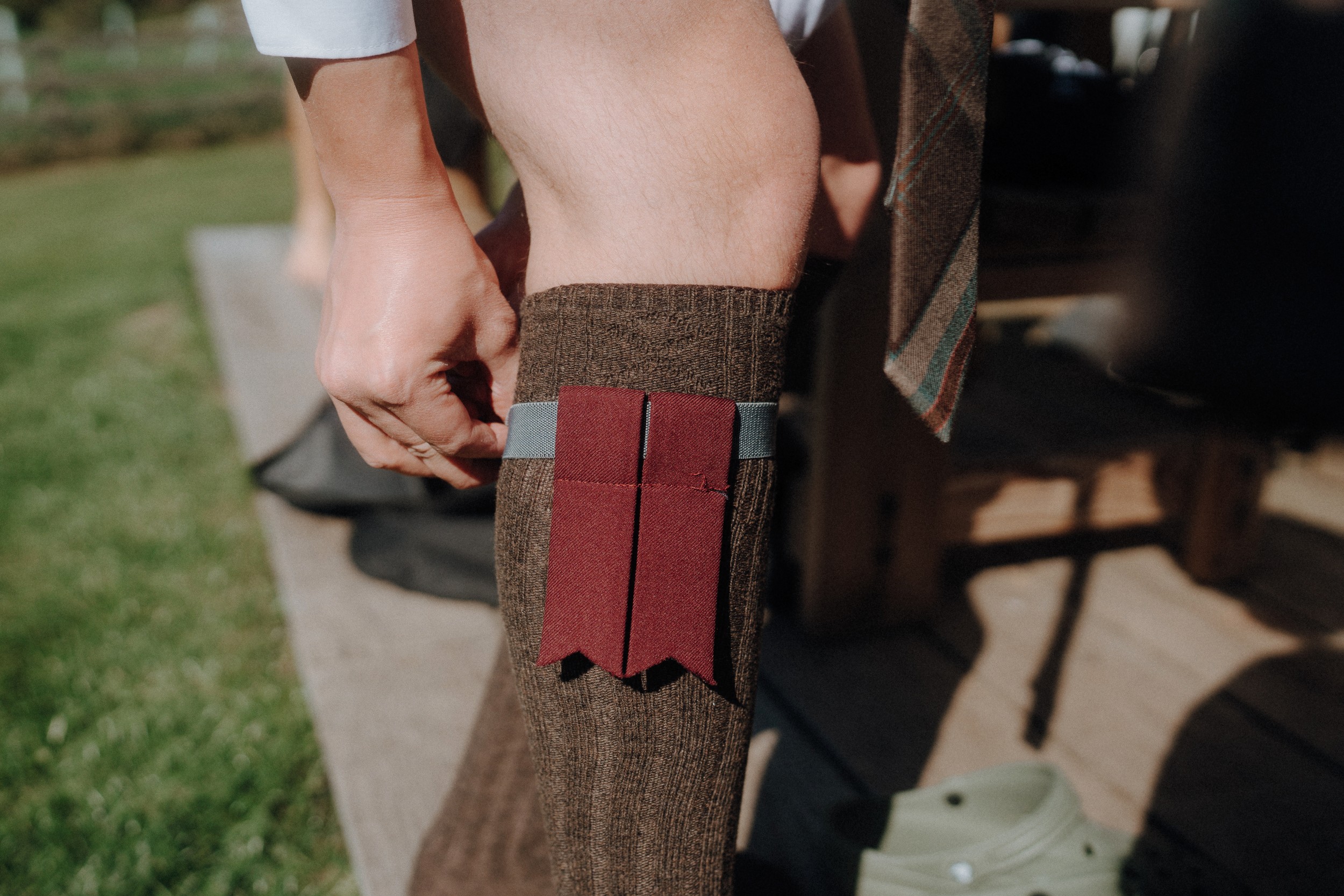 A person adjusts a maroon ribbon and gray band on a brown knee-high sock while standing outdoors on a wooden surface.