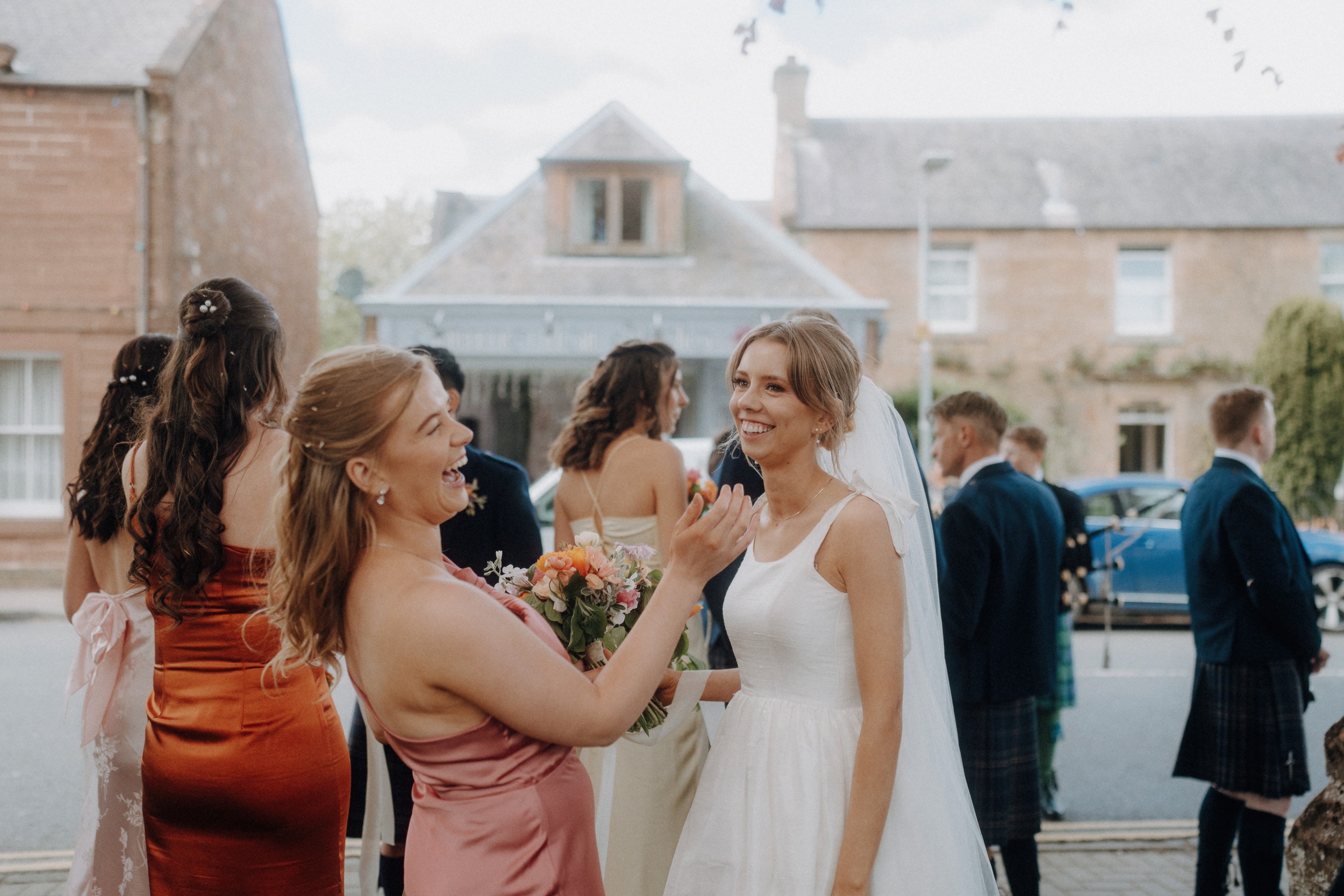 Two women, one in a white dress and veil and the other in a pink dress holding a bouquet, smile and interact outside with others nearby on a street.