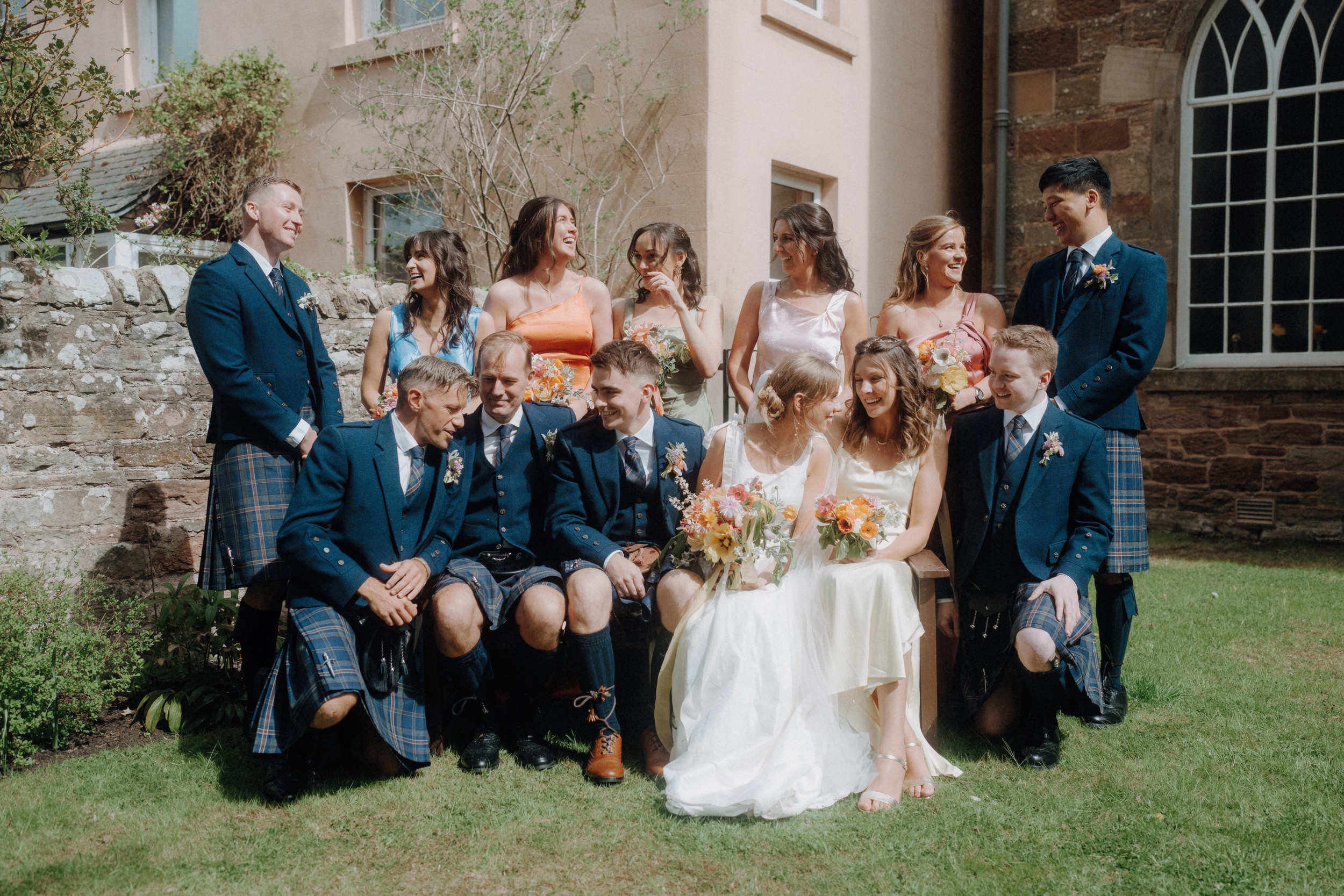 A wedding party poses outdoors, with men in kilts and jackets and women in pastel dresses, sitting and standing on a lawn in front of a stone building.