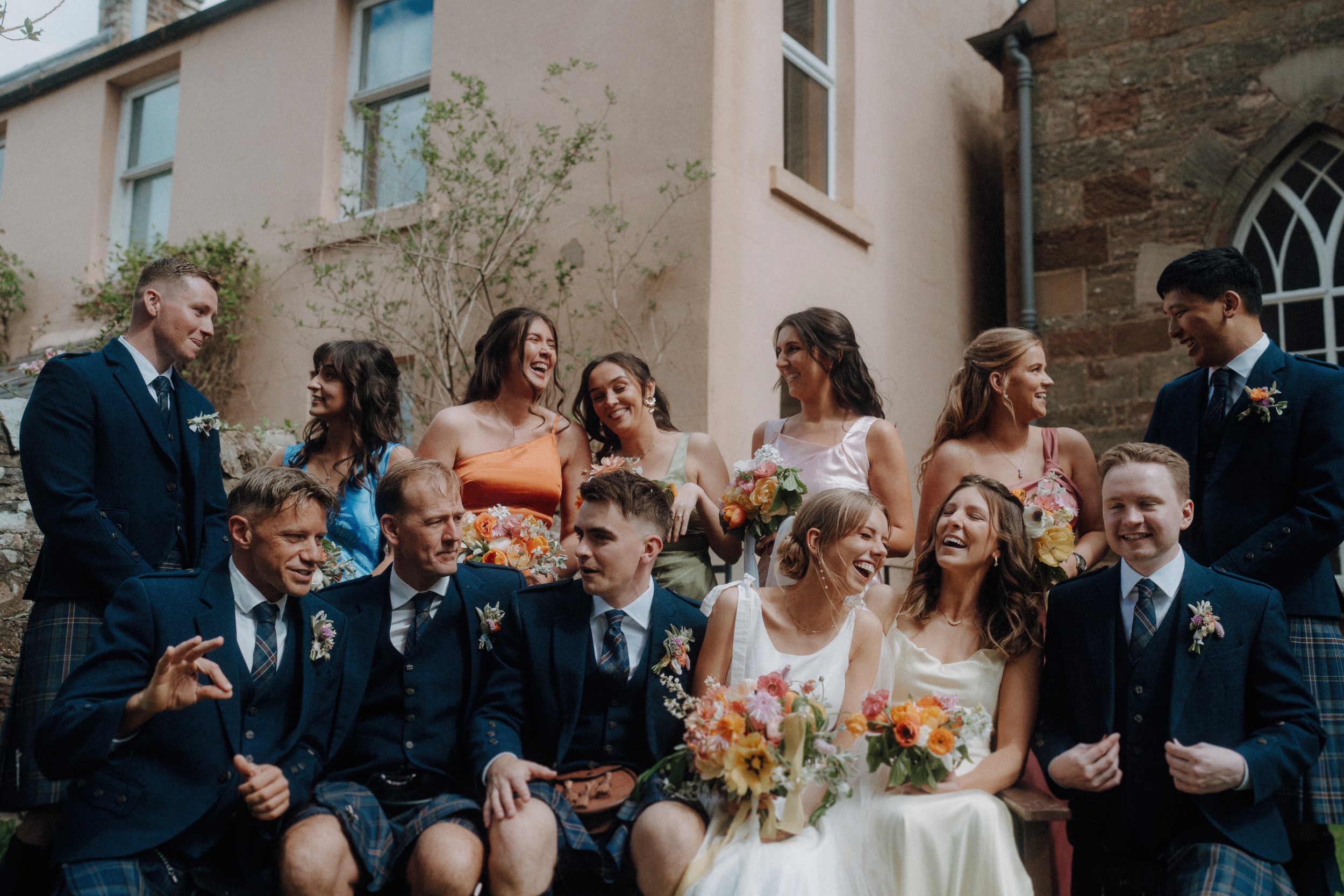 A wedding party poses outdoors, with seven people in suits and kilts and six people in colorful dresses, smiling and holding bouquets in front of a stone building.