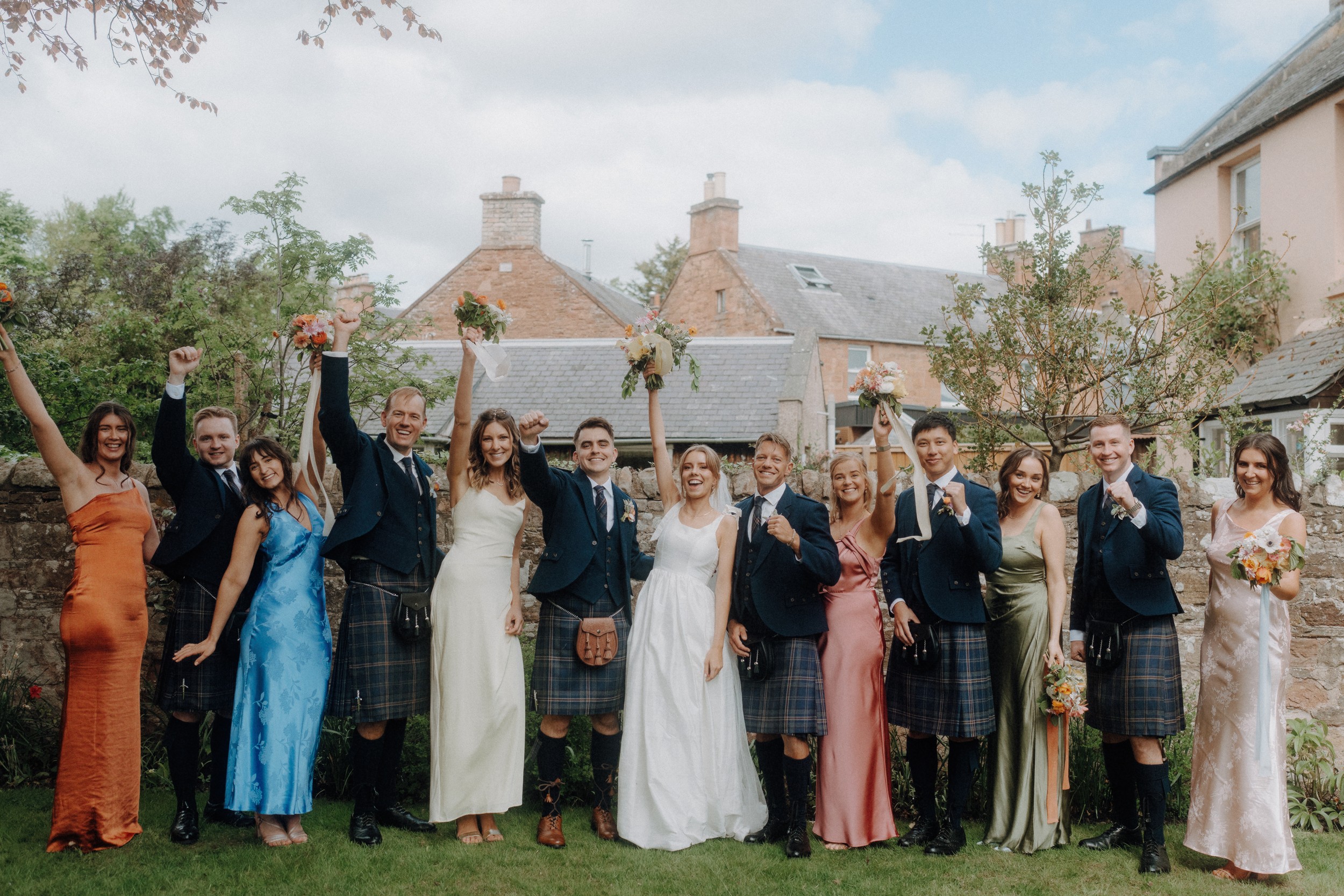 A wedding party poses outdoors, with men in kilts and women in dresses, holding bouquets and raising their arms in celebration.