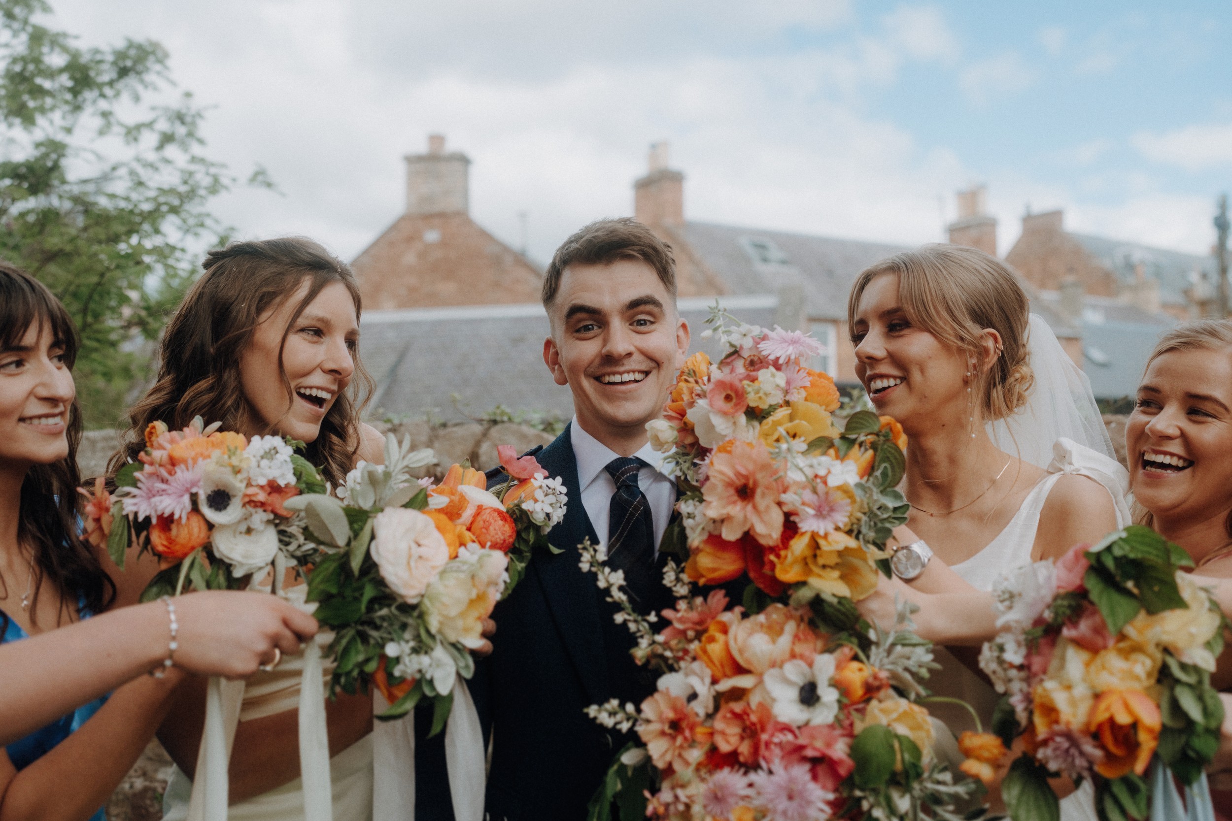 A group of five people dressed formally, holding colorful bouquets, smile together outdoors in front of stone buildings.